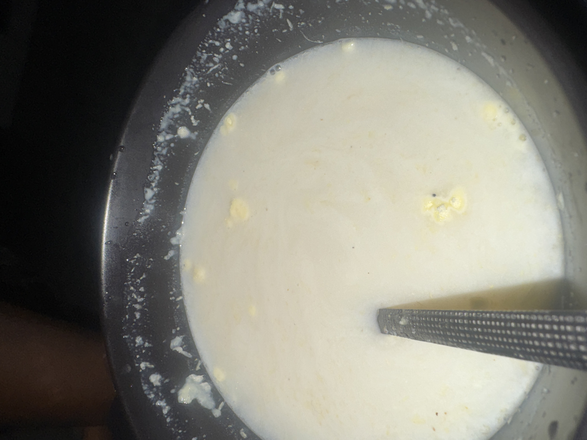 The image shows a close-up, overhead view of a dark, circular pot filled with a creamy, light-colored liquid, likely milk. Small, yellow clumps, possibly curdled milk or butterfat, are visible dispersed throughout the liquid and clinging to the rim of the pot. A metallic spoon, with a textured grip and a flat utensil end, rests submerged in the liquid, pointing towards the bottom of the pot. The rim of the pot also has dried white residue on its inner surface, indicating it has been used recently. The overall lighting is dim, with some reflections on the surface of the liquid. The background is dark and out of focus, suggesting the photograph was taken indoors. There is no discernible indication of the location in Abuja, Nigeria, within the image itself.