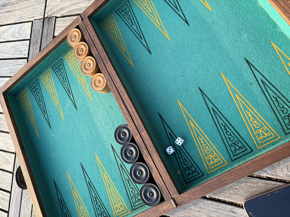 An elevated, downward-angled photograph captures an open backgammon board resting on weathered wooden decking outdoors in Bû, France. The board features a dark brown wooden frame surrounding a teal felt playing surface. The points are delineated by alternating black and yellow triangles, with the yellow points containing intricate scrollwork designs. Six black circular checkers are stacked along the left side of the central bar, closer to the bottom-left corner of the board's interior. Four light-colored circular checkers are stacked along the right side of the central bar, closer to the top-right corner of the board's interior. Two white dice rest on the teal felt in the bottom-left quadrant of the board's playing surface, with one die displaying a single pip and the other displaying two pips. The surrounding environment consists of visible wooden planks, indicating an outdoor setting, illuminated by natural daylight. No individuals are present in the image.