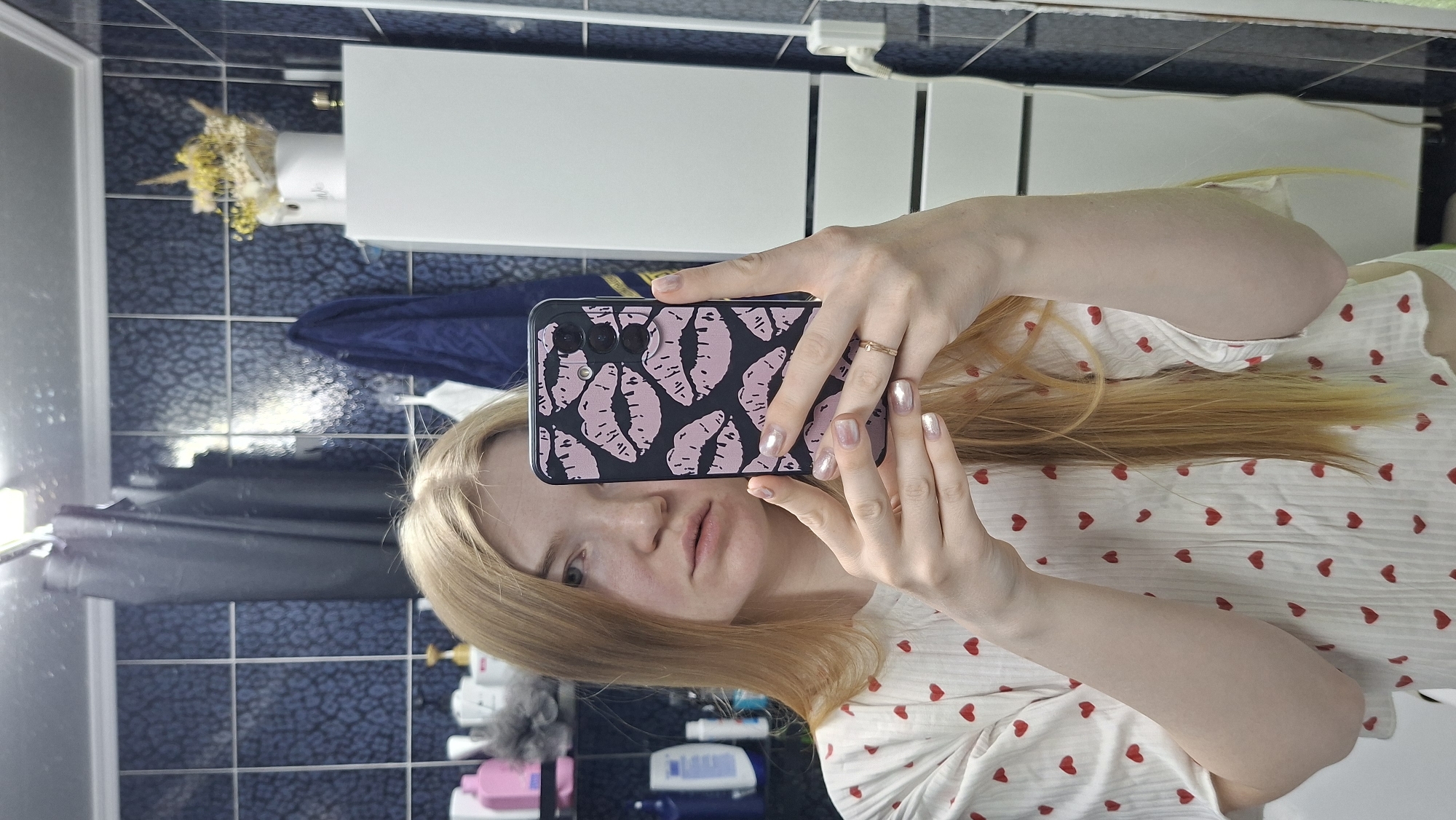 A young woman takes a mirror selfie in what appears to be a bathroom. She is holding a smartphone with a black case patterned with pink lip prints, obscuring part of her face. She has light skin and blonde hair. She wears a white pajama top with red hearts. The bathroom has blue tile walls and white fixtures, including a sink and a cabinet. The reflection in the mirror shows toiletries on a shelf. It is not possible to determine if this image was taken in the city of Chaikovsky, Russia. The image shows no location-specific details.