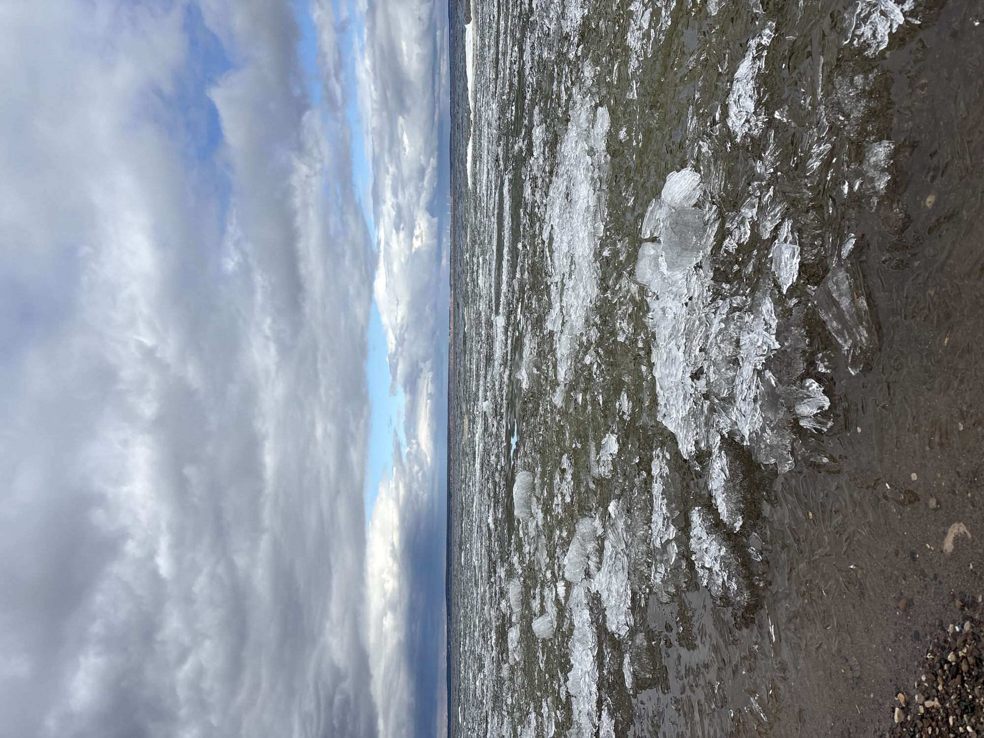 The image shows a body of water partially covered in ice, with a cloudy sky overhead. The ice appears to be broken into smaller pieces, close to the shore, where it meets a rocky or gravelly beach. In the distance, a dark line suggests the opposite shore or horizon. The overall impression is of a cold, overcast day at a shoreline with partial ice cover. While the location is presumed to be in the city of Ulyanovsk, Russia based on the prompt, there are no specific geographical landmarks or people in the image that confirm this.
