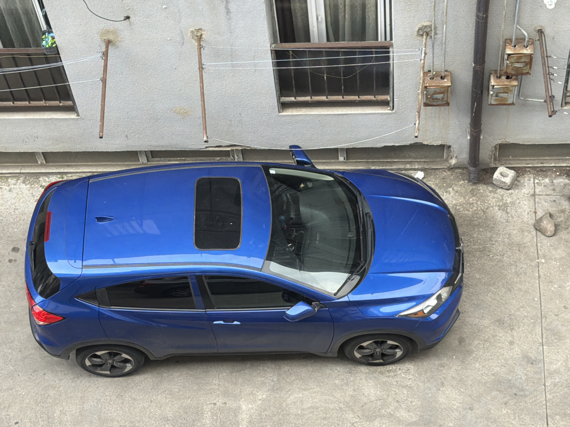 A blue SUV is parked on a concrete surface in what appears to be an urban alleyway or courtyard. The vehicle is viewed from directly above, showcasing its roof, including a sunroof. The car's body is reflective, capturing the surrounding environment, though the reflections are somewhat distorted. The scene is outdoors, with a gray building wall visible in the background, featuring several windows with metal bars and some rusty electrical boxes or conduits. A dark vertical pipe runs down the wall. The ground is made of rough concrete, with a few scattered pebbles and a small, light-colored concrete block. The lighting suggests it is daytime, possibly overcast, as there are no strong shadows. The overall atmosphere is utilitarian and somewhat aged, with the weathered building and outdoor parking area. No people are visible in the frame, and the car appears to be stationary. There is no visible text in the image.