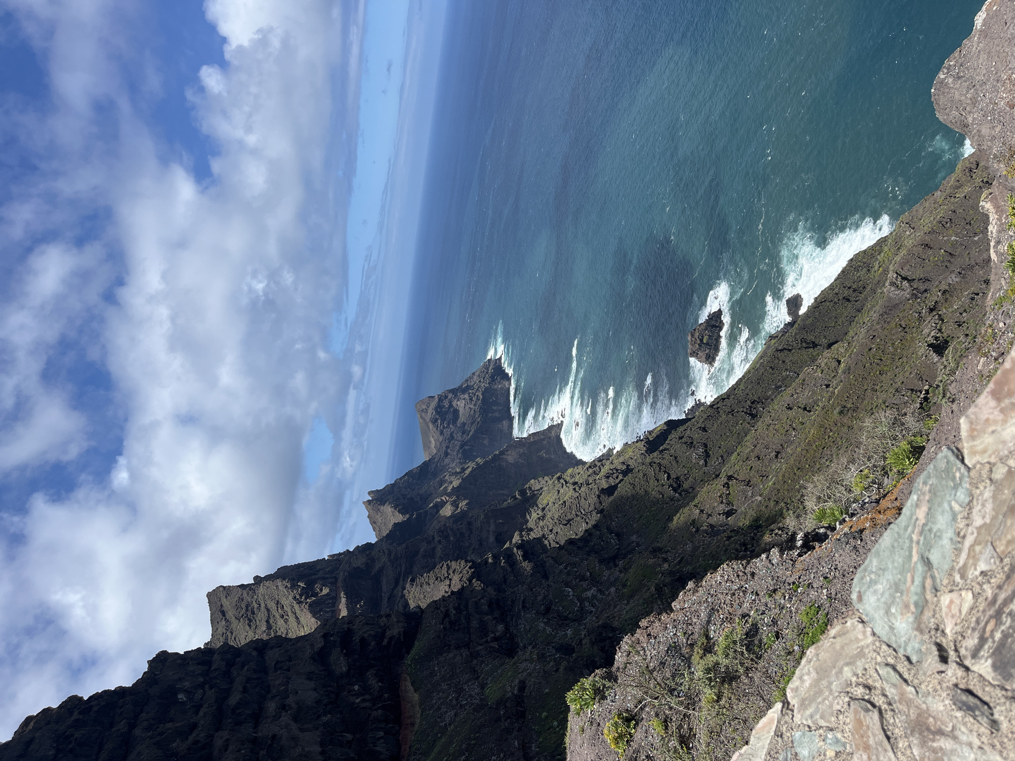 The image captures a dramatic view from a cliff overlooking the coastline of Agaete, Spain. The vantage point is high, providing a steep downward angle onto the dark, rugged cliffs that plunge into the turquoise ocean. White waves crash against the base of the cliffs and rocky outcrops. Above, a blue sky is interspersed with patches of white clouds. The foreground features stone and rocky terrain with sparse vegetation. The photo emphasizes the ruggedness of the terrain, contrasting the rough cliffs with the sea and sky.