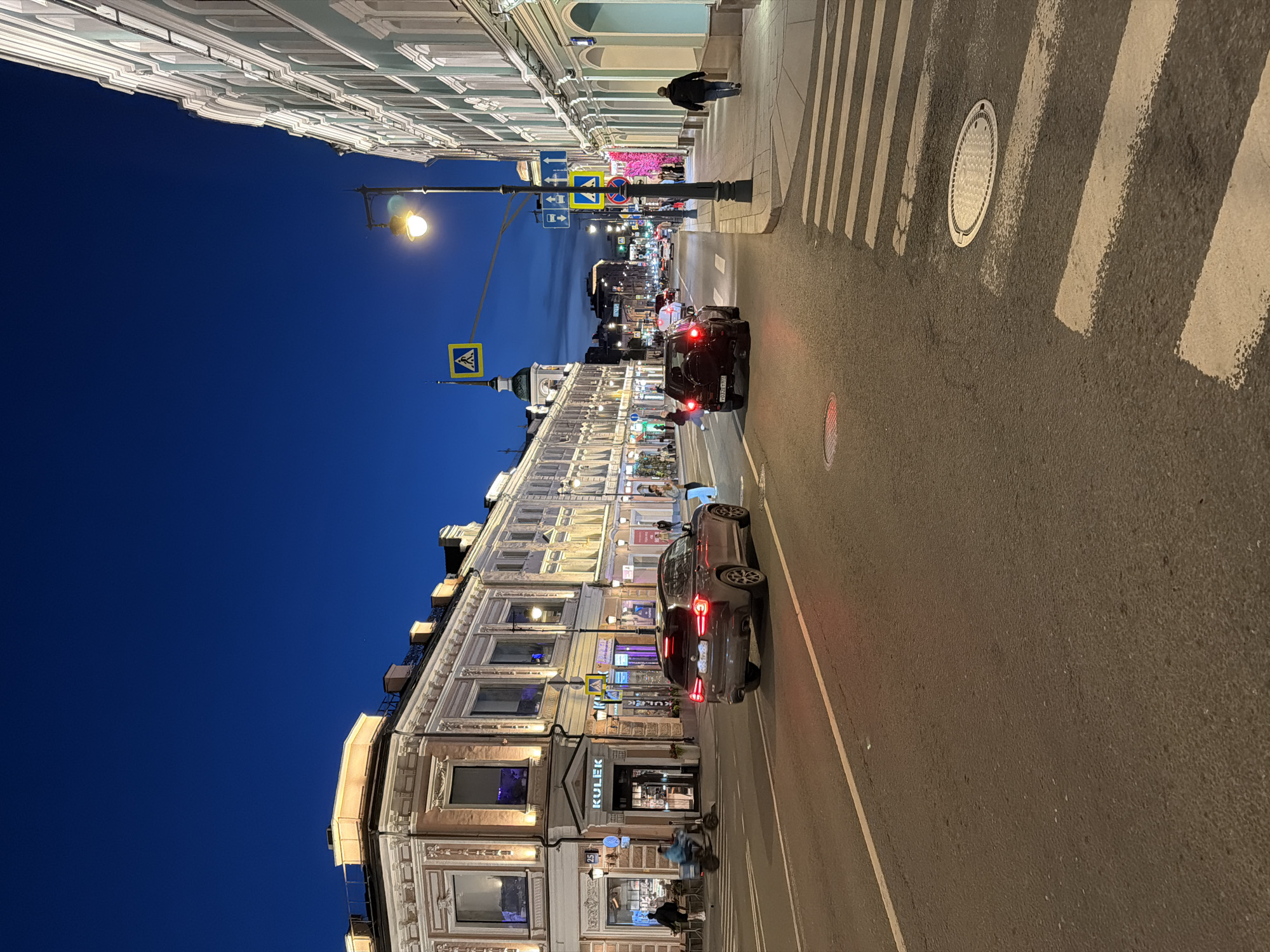 The image, correctly oriented, captures a vibrant city street scene at dusk or night. Ornate, multi-story buildings with classical architectural details, such as cornices and decorative window frames, line the left side of the street. These buildings are brightly illuminated by warm artificial light spilling from their windows and ground-floor shops. A deep blue sky, indicating early evening, stretches above.
On the right, an asphalt road features a prominent pedestrian crossing marked with white zebra stripes, and at least two manhole covers are visible within the road surface. Two cars are present on the street; a dark SUV with illuminated red taillights is in the foreground, facing away from the viewer, and another vehicle is further down the road.
Several pedestrians are seen on the wide sidewalks. One person is distinctly walking on the upper sidewalk, and others are visible near the brightly lit shop entrances. Streetlights, some emitting a bright yellow glow, are mounted on poles along the street, contributing to the overall illumination. Various traffic signs are scattered throughout the scene, including yellow diamond-shaped pedestrian crossing signs, blue rectangular signs with white directional arrows, and a red circular "no stopping" or "no parking" sign.
Visible text includes "KULEK" on a shop sign, with the number "25" directly below it. The license plate on the dark SUV has partially discernible characters, including "Y822", which appear to be part of a Cyrillic script plate. The scene conveys a lively urban evening atmosphere.