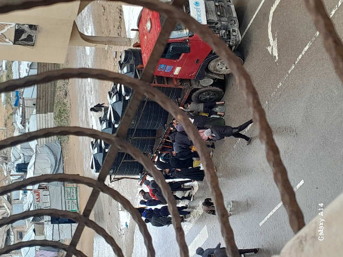 The image captures an elevated view through an ornate, reddish-brown metal grate, overlooking a street and a settlement in Khan Yunis, Palestinian Territory. On a wet, asphalt road with visible white crosswalk markings and a turn arrow, a red heavy-duty truck is parked. The truck's cab displays a "unicef" logo and the number "528." Its bed carries at least eight large, black, cylindrical water tanks. A group of approximately 20-30 individuals, mostly dressed in dark, heavy clothing, are gathered around the rear and side of the truck. Several people are bending low, appearing to fill yellow and white plastic jerrycans and buckets from the truck. One person is walking across the crosswalk with a white bucket, while another individual is bent over near a white container on the wet pavement. In the background to the left and center, a dense collection of makeshift shelters and tents, constructed from white and grey tarpaulins and corrugated metal, indicates a camp. Arabic script is visible on some structures, including "المجد" (Al-Majd). To the right, a beige, multi-story building with arched, decorative windows is visible. The ground is wet throughout, suggesting recent rain.