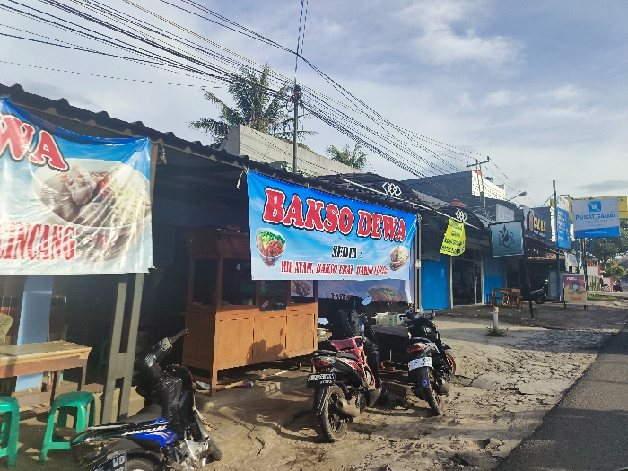 The image shows a street scene in Cikalong, Indonesia (latitude: -6.748, longitude: 107.446). The scene features a street lined with small shops and businesses. On the left side, a food stall with a sign that reads "Bakso Dewa" is visible. The sign advertises "Mie Ayam, Bakso Daging, Bakso Ikan." There are two motorcycles parked outside the stall. A blue and white awning is visible above the stall, with a sign that says "Wa" in large letters above the word "Lencang." A small wooden table and two green plastic chairs are also visible in front of the stall. The right side of the street features a shop with a blue awning. A sign with the word "Kul" is visible above the shop door. There are several electrical wires running overhead across the street. The street appears to be cobblestone. The image shows a bright, sunny day with a blue sky with a few clouds.