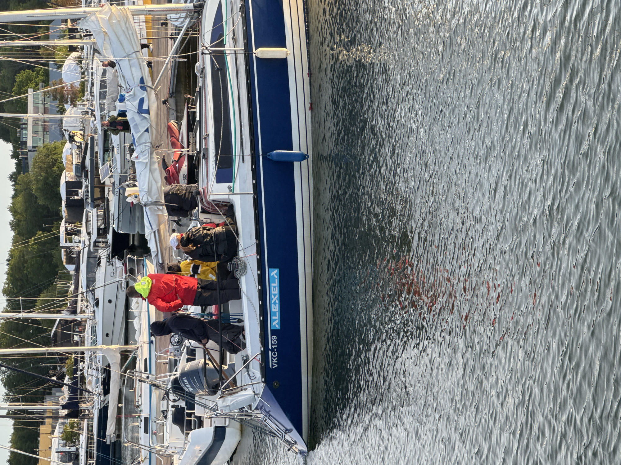 The image shows a group of people standing on the deck of a blue and white sailboat docked in a marina. The boat has the registration "VKC-159 ALEXELA" printed on its side. In the background, other sailboats and buildings are visible, suggesting an urban waterfront setting. The water is calm with ripples reflecting the sunlight. One person on the boat is wearing a bright yellow jacket. The scene appears to be a sunny day, possibly in the late afternoon given the lighting. The location is the city of Tallinn, Estonia.