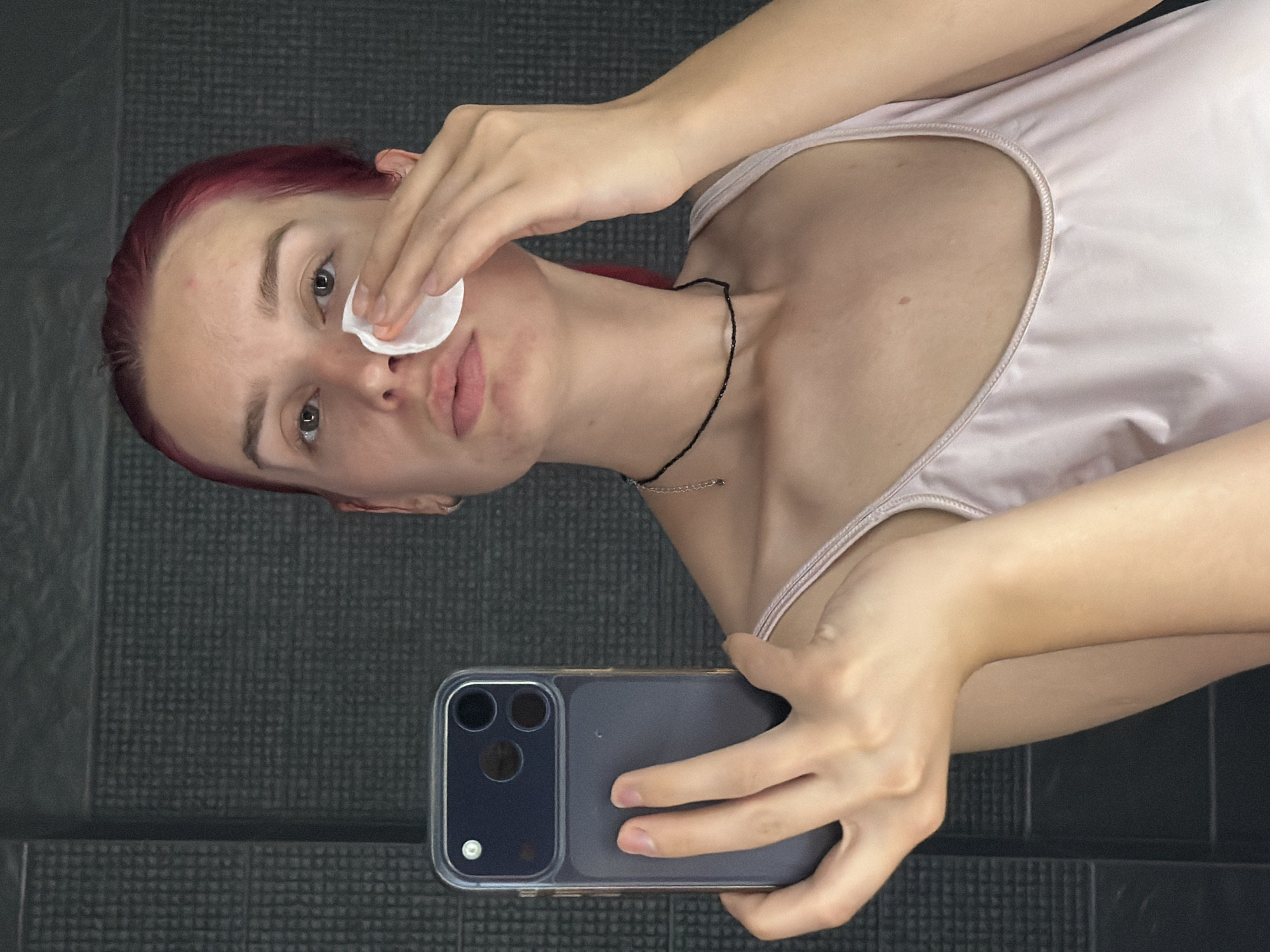 A young woman with vibrant red hair is shown in a mirror selfie. She is holding a dark-colored smartphone in her left hand, its camera facing her. In her right hand, she holds a white cotton pad, which she is using to cleanse her face. She is wearing a light pink camisole. The background appears to be a tiled wall with a dark gray, textured pattern. The scene is indoors, likely a bathroom or bedroom, and it's not possible to determine the specific location within the city of Chaikovsky, Russia, from the visual information provided.