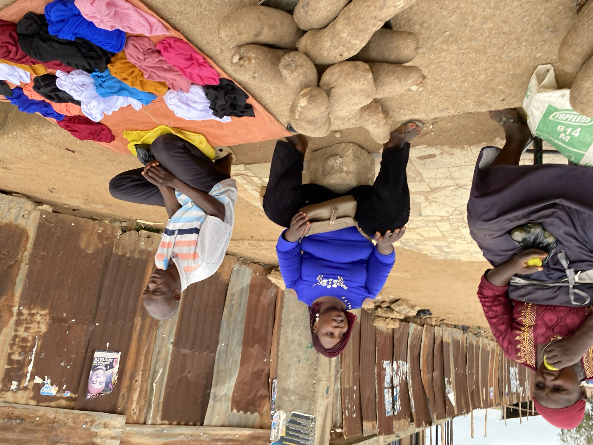 The image captures an outdoor market scene in Bukuru, Nigeria, featuring three individuals amidst various goods for sale. The setting is characterized by a dirt or packed earth ground and a background of rusty, corrugated metal sheets forming makeshift stalls or walls.
To the left, a man wearing a white, blue, and orange striped short-sleeved shirt and dark trousers is seated on the ground with his legs crossed, looking away. Next to him, an array of folded clothes in diverse colors including pink, blue, black, white, red, yellow, and brown, is neatly displayed on an orange mat or tarp, suggesting they are for sale.
In the center of the frame, a woman dressed in a royal blue long-sleeved top with white embroidery and dark pleated trousers is seated on a low stool. She wears a reddish headscarf and holds a light-colored handbag in her lap, her gaze directed towards the viewer.
To the right, another woman, wearing a dark purple/grey dress over a red patterned shirt and a reddish-pink headscarf, is actively eating a light green fruit. She is positioned near a substantial pile of large, irregularly shaped yams, also laid out on the ground, indicating they are offered for sale. A green sack bearing the text "TOPFEEDS" and "914 M" is visible near the yams.
The scene appears to take place during daylight hours, suggesting clear weather conditions suitable for market activities. On the corrugated metal wall to the far left, a partially torn flyer or poster is affixed, with the word "APUTAZE" and phrases such as "NOW I'M DECLARED" faintly legible alongside a depicted face. The overall mood is casual, with the individuals engaged in market life without strong emotional expressions.