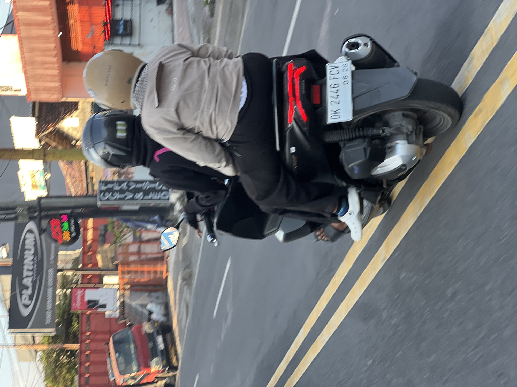 Two individuals, wearing helmets and riding a motorcycle, are captured in motion on a road in Denpasar, Indonesia. The person in front wears a black jacket with a pink strap visible, while the person behind is in a tan jacket. The motorcycle's rear light is illuminated, and a license plate reading "DK 2446 FCV 06-28" is clearly visible. The road features double yellow lines, indicating traffic lanes. In the background, a red truck is parked near a building with a "PLATINUM" sign. The overall scene suggests a typical urban street environment in Denpasar during daylight.