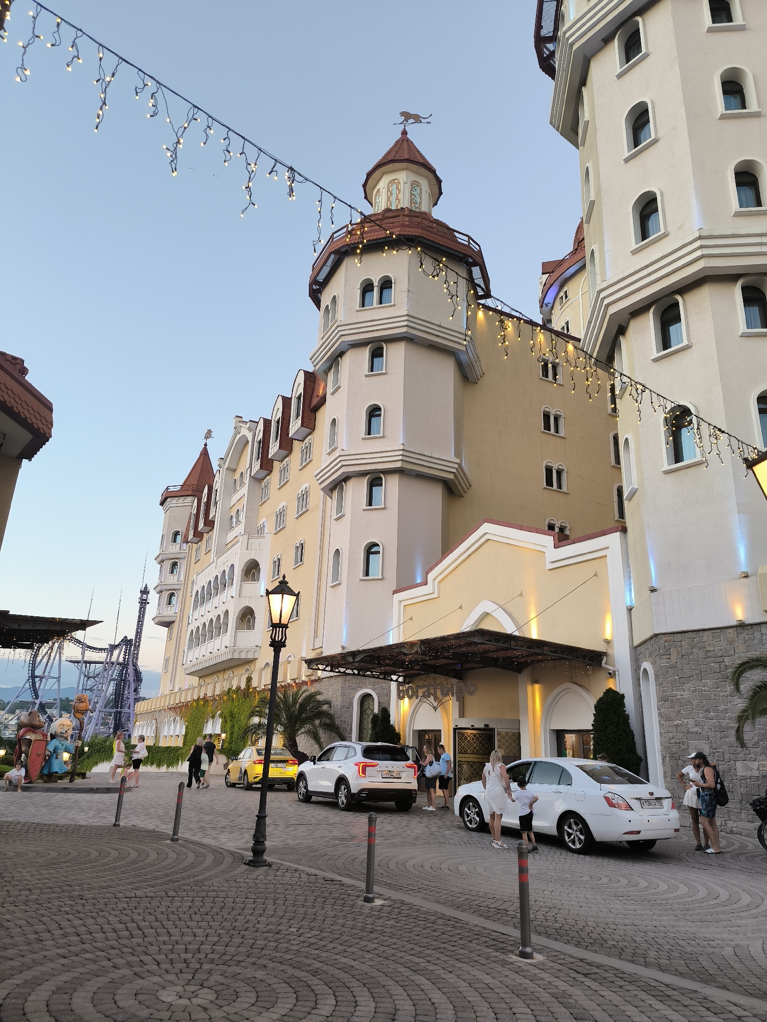 This is a medium-shot, eye-level, outdoor photograph of the complex of buildings designed in a fairytale castle style, located in the city of Sirius, Russia. The buildings are tall and colorful, with numerous towers, arched windows, and red-tiled roofs. String lights with small, bright bulbs are draped across the scene, adding a festive atmosphere. In the foreground, a cobblestone walkway leads to the entrance of one of the buildings, where people are seen arriving and departing. Several cars are parked on the side of the road, including a yellow sedan and a white SUV. Behind the buildings, a roller coaster track is visible, indicating the presence of an amusement park or entertainment venue. The sky is clear and blue, suggesting it is daytime. The overall impression is that of a lively and enchanting tourist destination.