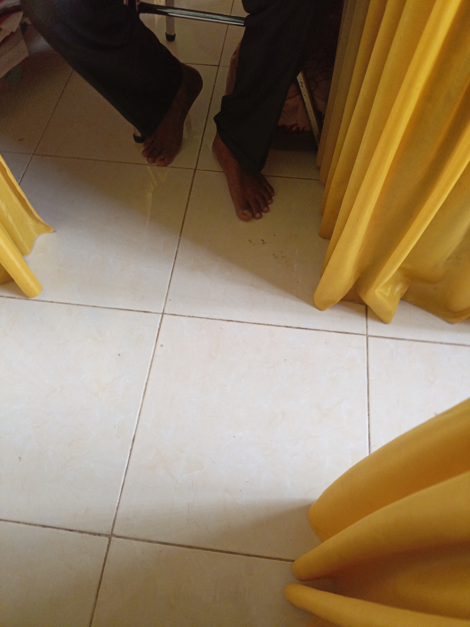 The image shows the lower legs and feet of two individuals standing on a tiled floor. The person on the left is wearing dark pants and is barefoot. Their left foot is visible, showing toes. The person on the right also appears to be wearing dark pants and is barefoot, with their right foot partially visible. There is a large expanse of yellow fabric to the right, which appears to be a draped curtain or tablecloth, and another piece of yellow fabric in the bottom right corner. The environment suggests an indoor setting with a tiled floor, possibly a home or a public space. There is no discernible information to pinpoint the exact location within Lhokseumawe, Indonesia, based solely on the visual content of the image.
