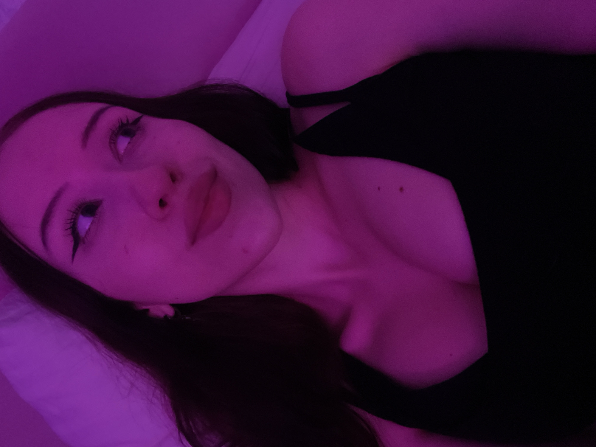 The media file is a close-up, low-angle selfie of a young woman lying down. She has long, dark hair and is wearing a black top. The scene is lit with purple and pink ambient light, creating a moody and intimate atmosphere. Her face is tilted upwards and to the left, with her eyes looking in that direction. She has noticeable eyeliner and mascara on, accentuating her eyes. Her lips are slightly parted. Her skin appears smooth and is reflecting the colored light. A portion of her collarbone and chest are visible. The background is a soft, blurred texture, likely a pillow or bedsheet, illuminated by the same purple light. The overall impression is one of relaxation and personal reflection, possibly taken in a bedroom setting at night. There is no visible text in the image.