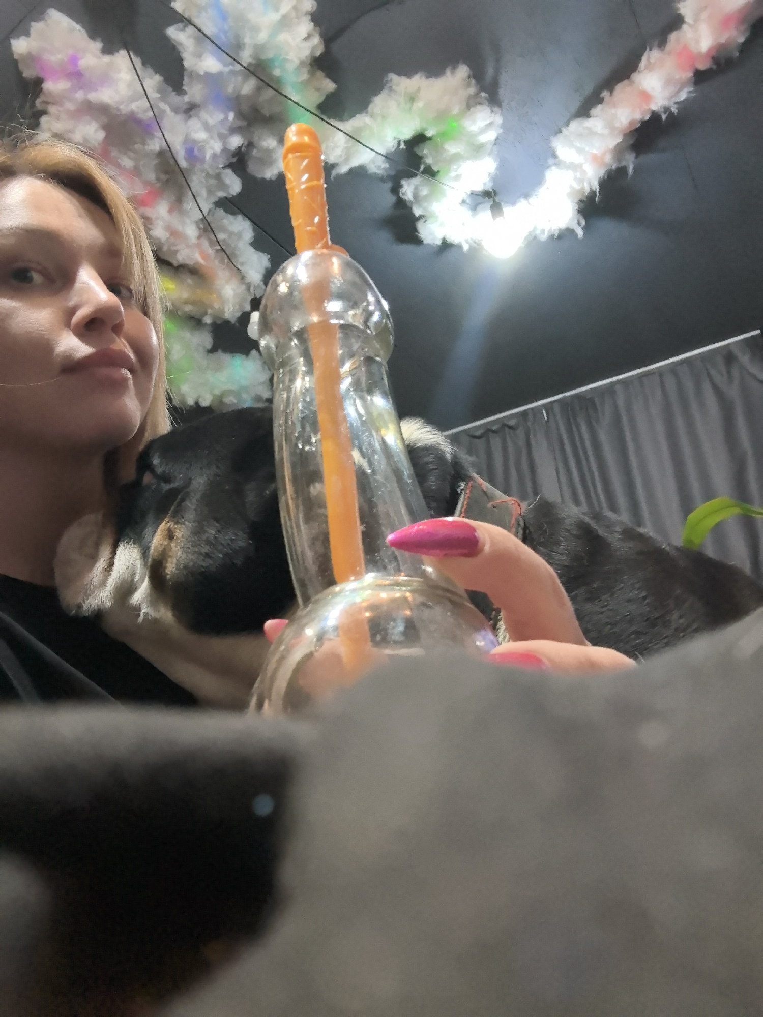 The image depicts an indoor scene in Noviy, Russia, featuring a woman and her dog. A woman with fair skin and light brown hair, wearing a black top, is partially visible on the left. She holds a clear glass beverage container with a distinctive orange, phallic-shaped straw. Her hand is adorned with bright pink nail polish. She displays a subtle, content expression.
A black and white dog is nestled against the woman's side, resting its head near her chest, indicating a close and comfortable bond.
The background features a dark ceiling decorated with fluffy white material resembling clouds, illuminated by multicolored lights (green, red, white) and a visible light bulb. This unique decor suggests a themed establishment like a bar or cafe. A dark curtain or backdrop runs across the upper right side of the image, and a small green plant is partially visible on the far right. The foreground is partially obscured by dark fabric. The scene conveys a relaxed and casual atmosphere, likely taking place in the evening or a dimly lit indoor setting. No text is visible in the image.