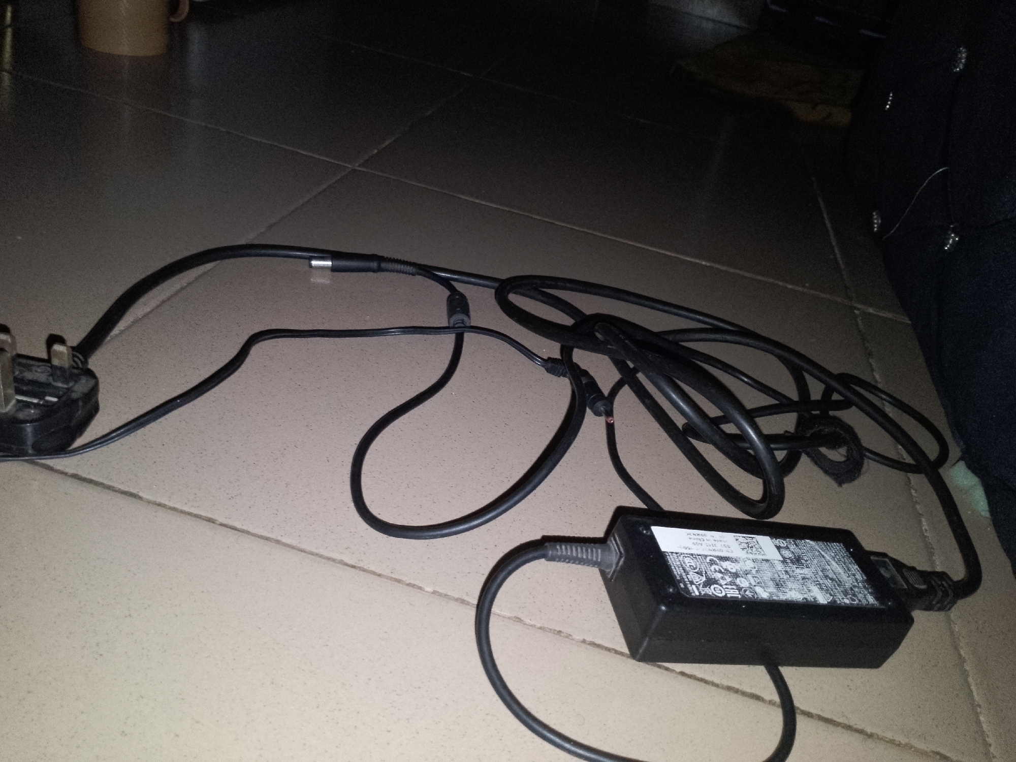 The image shows a close-up, overhead view of a black power adapter with its associated cables lying on a tiled floor. The power adapter is rectangular and black, with a white label on top displaying text and symbols, some of which are obscured or illegible. One end of the adapter has a black cable emerging from it, which then loops downwards. The other end of the adapter has a plug with three rectangular prongs, likely a UK-style plug, attached to another black cable. This cable runs upwards and then loops back towards the adapter, forming a tangled mass of wires. A thin metal connector is visible at the end of one of the cables. The floor is made of beige, square tiles with visible grout lines. In the background, there is a dark, out-of-focus area, possibly furniture or a wall, and a small portion of a light-colored mug is visible in the upper left corner. The lighting in the image is dim. There are no people or animals present in the scene. The image is taken in the city of Jauro Yinu, Nigeria.