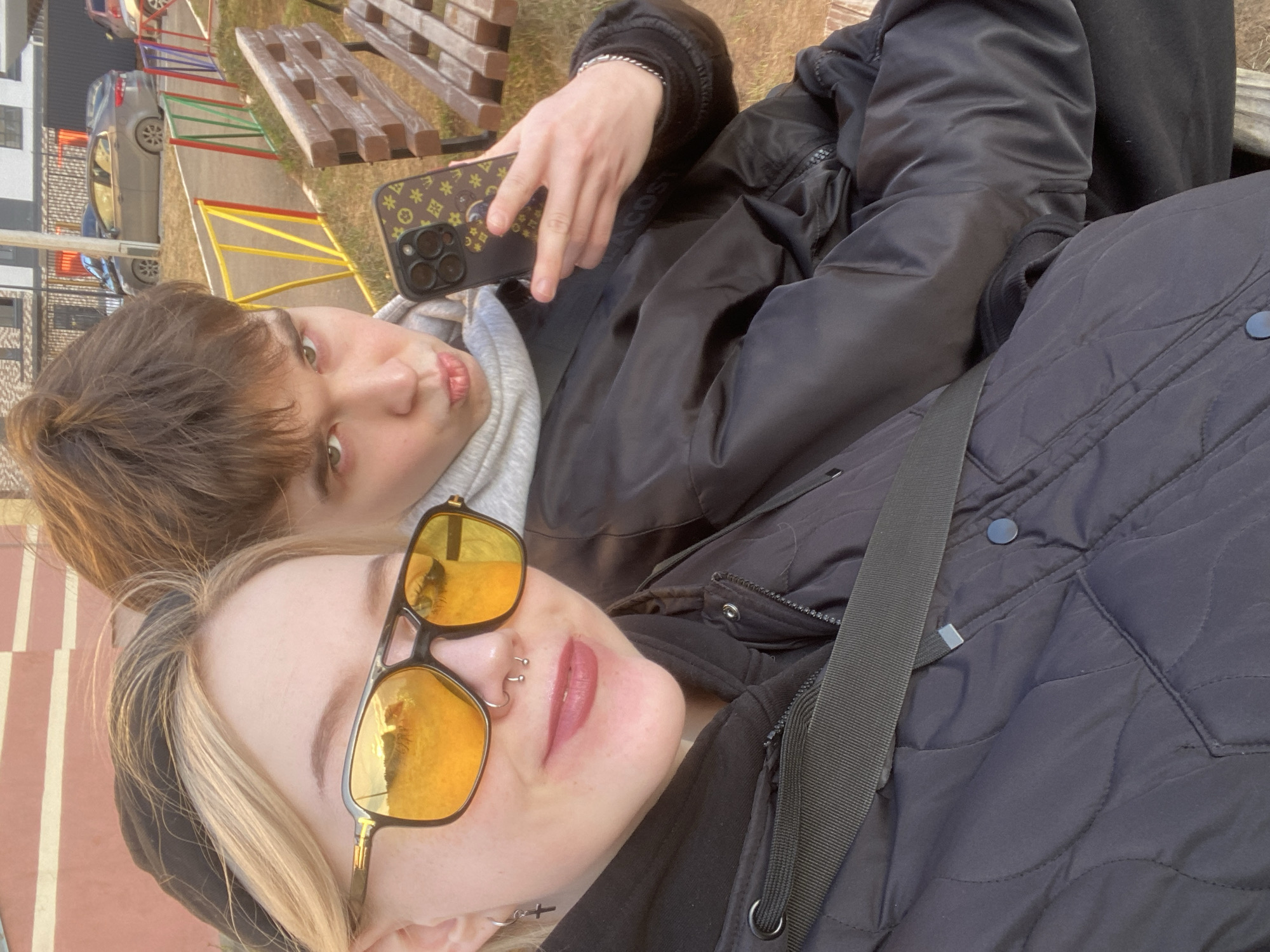 Two individuals are depicted in a selfie-style photograph. The person in the foreground is wearing yellow-tinted sunglasses, a black headband, and has multiple piercings on their nose. They are wearing a black quilted jacket with a visible black strap across it. The other person, positioned slightly behind and to the side, holds a phone with a patterned case. They are wearing a grey hooded sweatshirt and a black jacket. The background shows a playground with colored railings, a wooden bench, cars, and a building. Based on visual landmarks, the location appears to be in the city of Izhevsk, Russia.