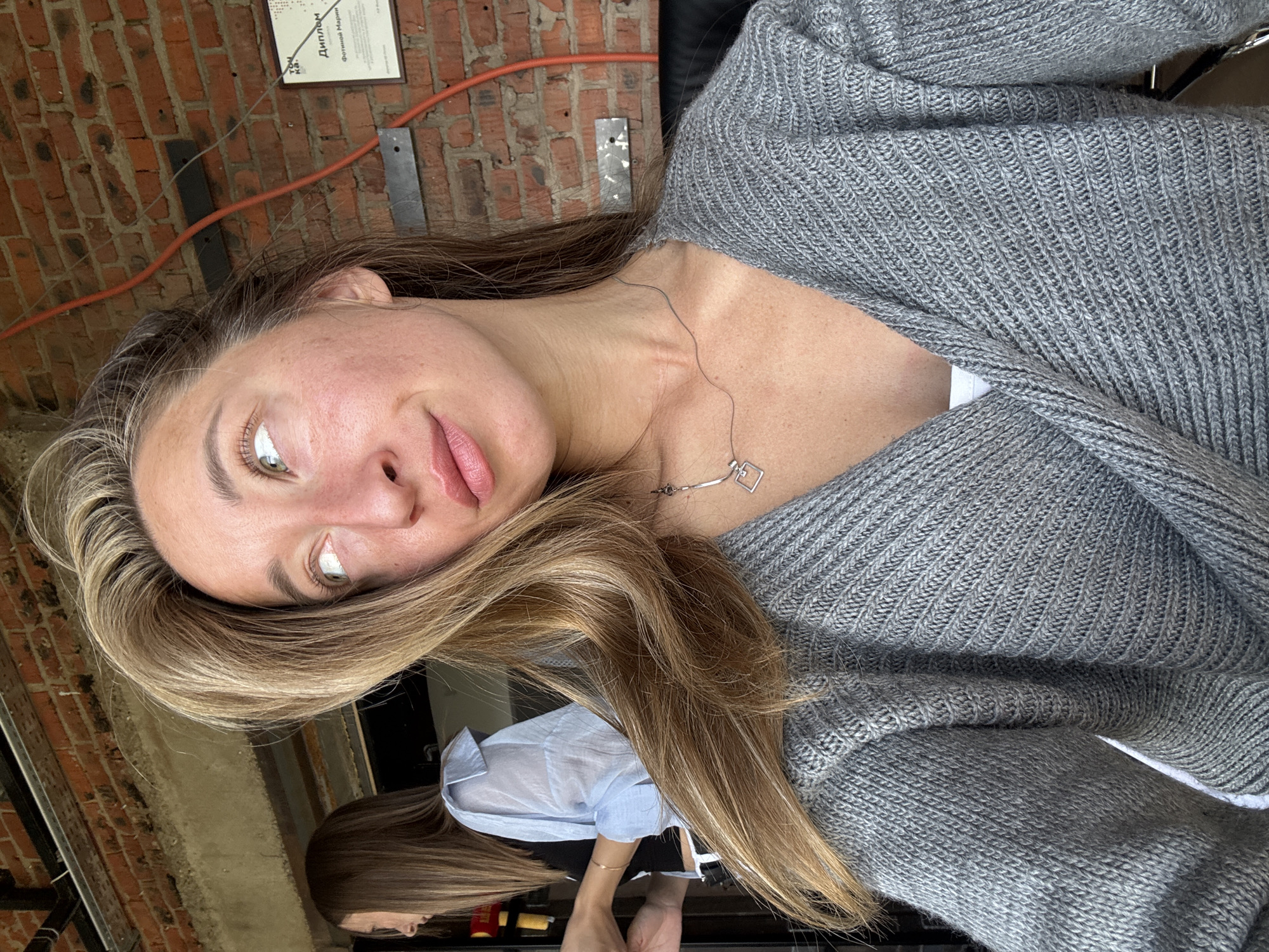 The image shows a close-up of a woman with blonde hair and green eyes, looking to her left. She is wearing a grey knitted cardigan and a white top. In the background, out of focus, another woman with brown hair is visible. The setting appears to be indoors, with a brick wall behind the subjects. A framed certificate or diploma is hanging on the wall. The overall impression is that of a casual, candid moment captured at a workplace or a communal space. There is no direct indication of the city of Chaikovsky, Russia, but the certificate has Russian text, suggesting a Russian-speaking environment.