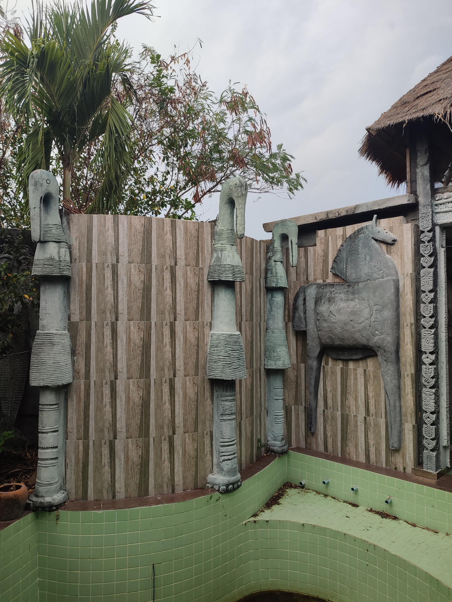 The image shows an empty, green-tiled pool in an outdoor setting. Three abstract, gray, stone sculptures with cylindrical segments and head-like tops stand along the pool's edge against a wooden fence. A relief sculpture of a horse is mounted on the fence. A thatched roof structure is visible in the upper right corner. The sky is overcast. Tall palm trees and other vegetation are in the background. Based on prior knowledge of the location, the scene is likely located in Pattaya City, Thailand. There are no people visible in the image.