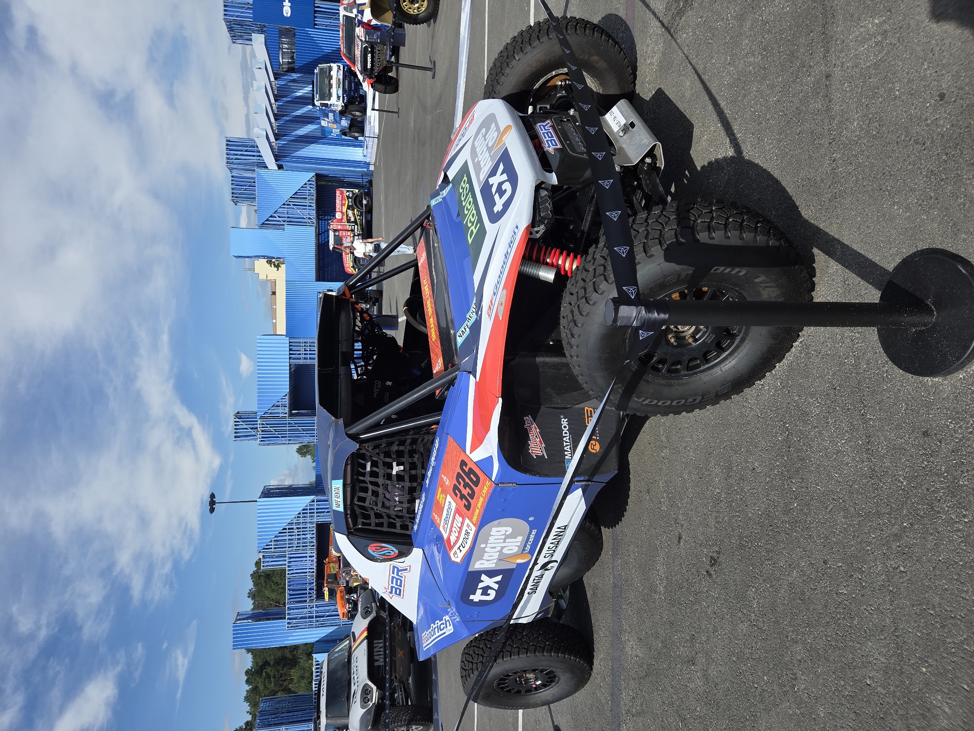This image depicts a rally car, likely a buggy or Trophy Truck, on display outdoors in the city of Yekaterinburg, Russia. The vehicle is predominantly blue and white with red accents, featuring numerous sponsor decals including "tx Racing Oil," "BFGoodrich," and "Motul." The car has large off-road tires and a roll cage visible. In the background, there are blue modular structures, possibly part of a temporary exhibition or event setup, along with other vehicles including a white MINI-branded car, several trucks, and a Toyota vehicle. The sky is blue with scattered clouds, and the ground appears to be asphalt. A stanchion with a black ribbon featuring white triangular emblems is in the foreground, indicating a barrier for spectators. The lighting suggests it is daytime.