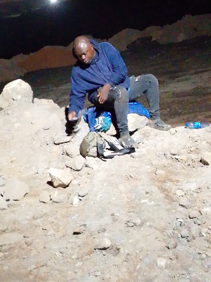 A dark-skinned man sits atop a rocky mound, wearing a blue hoodie and dark jeans. He looks down, holding something in his left hand. Behind him is a blue patterned cloth. To his right, on the rocks, is a clear plastic water bottle. A tan bag with black straps rests near his feet. The setting appears to be outdoors at night, with dimly lit rocky terrain surrounding him.