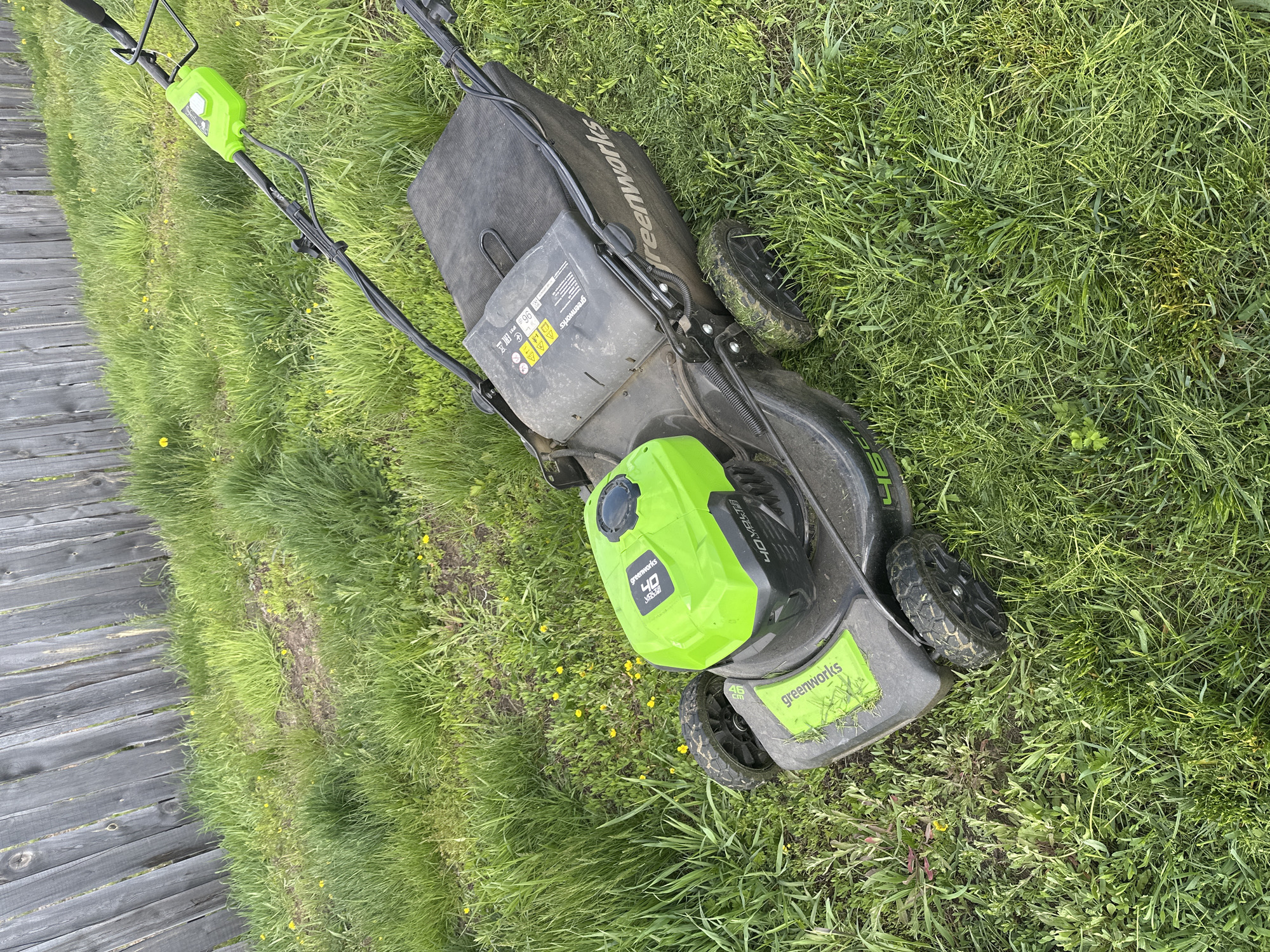 An overhead, slightly tilted shot captures a Greenworks lawnmower resting on overgrown grass next to a rustic wooden fence in what appears to be a yard or garden in Kuda, Russia. The scene is outdoors under an overcast sky, indicated by the soft, diffused lighting and lack of harsh shadows.
The primary subject is a black and bright green battery-powered Greenworks lawnmower. Its main body is black with a large lime-green battery cover or engine housing clearly visible towards the front. The grass is long and uneven, with scattered patches of small yellow wildflowers, possibly dandelions, particularly visible in the foreground and near the fence. Some areas of the grass appear slightly matted down, suggesting movement of the mower, or perhaps just uneven growth. The mower's wheels are dark and show signs of use, with some dirt and grass clinging to them.
Visible text on the mower includes "Greenworks" logos on the grass bag, the front deck, and the battery housing. The battery housing also displays "40V" and "Brushless," indicating its power source and motor type. Several warning symbols are present, including those for ear protection, foot protection, keeping bystanders away, and a recycling symbol. The noise level "96 dB" is also marked. The cutting width "46 cm" is visible on the front of the mower deck.
No people are visible in the image. The wooden fence, composed of weathered gray planks, runs along the left side of the frame, providing a boundary to the grassy area. The overall impression is one of a typical yard chore awaiting completion or having just started.