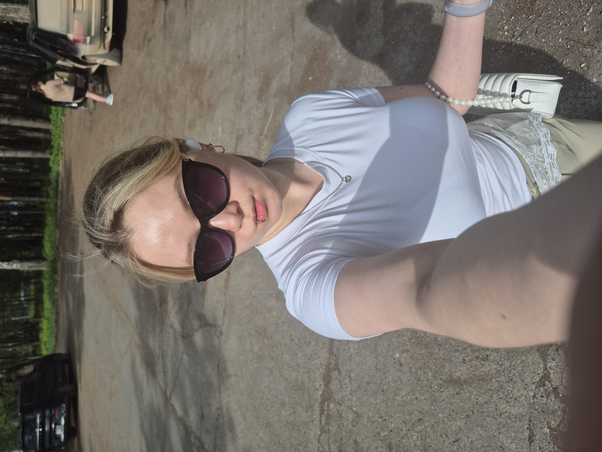 A woman with blonde hair and sunglasses is taking a selfie. She is wearing a white t-shirt, a silver necklace, and has a pearl bracelet on her wrist. A white purse is partially visible in the upper right corner of the frame. The woman is standing on a paved surface, likely asphalt, with a wooded area in the background. In the background, there is a dirt road and the back of a vehicle. The location could be Chaikovsky, Russia, based on the presence of the wooded background and the type of vehicles.
