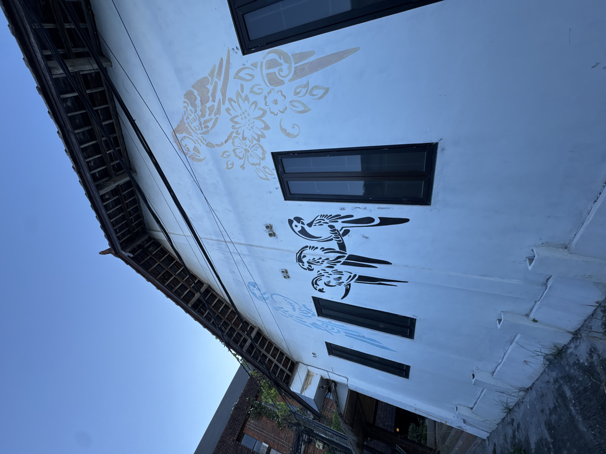 A low-angle shot features a white, multi-story building adorned with stylized bird and flower murals in Kuta, Indonesia. The building's exterior walls are painted with these decorative elements. Rectangular windows with dark frames punctuate the facade. Power lines stretch across the scene, partially obscuring the view of the building and contrasting with the clear blue sky visible in the background. The roofline of the building is visible, with a dark, overhanging eave. The image also captures a glimpse of adjacent buildings and greenery below, suggesting an urban setting.