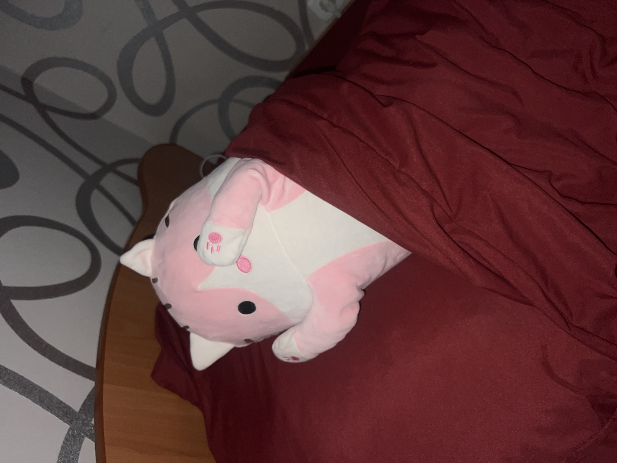 An indoor scene in Kazan, Russia, displays a large, pink and white plush stuffed animal tucked into a bed. The stuffed animal, possibly a fox or cat, features black eyes and nose, small pink dots on its cheeks, and white ears with pink inner details. Its paws, one resting on its head, show distinct pink pads and small pink markings. The creature is partially covered by a dark red duvet, with an underlying dark red fitted sheet visible. A light brown wooden bed frame or headboard is present on the left. The wall behind the bed has a light grey or white wallpaper adorned with a repeating pattern of dark grey, swirling, abstract lines. The lighting is dim, suggesting the image was taken in the evening or at night. No people are visible, and there is no discernible text. The overall atmosphere conveys coziness and comfort.