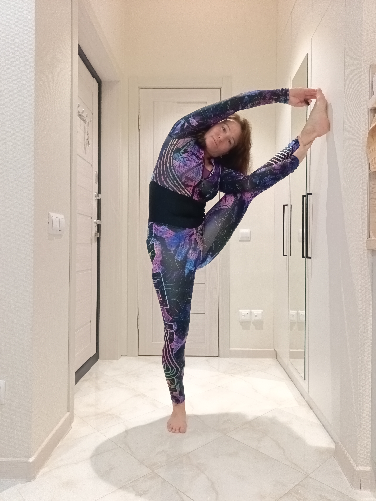 A woman in a colorful full-body athletic suit is performing a yoga pose. She is standing on her right leg and stretching her left leg upwards, with her foot resting against a white wall. Her left arm is extended upwards, touching her left foot, while her right arm is bent and extended forward. Her body is curved, and her head is turned towards the camera, with a neutral expression.
The background appears to be an indoor hallway or entryway. To the left, there are two white doors with silver handles and a white light switch. To the right, there is a tall white wardrobe with two black handles. The floor is tiled with a marble-like pattern. The lighting is bright and uniform, suggesting an indoor setting. The overall scene suggests a domestic environment, possibly a home or a gym.
This scene is not located in Sochi, Russia.