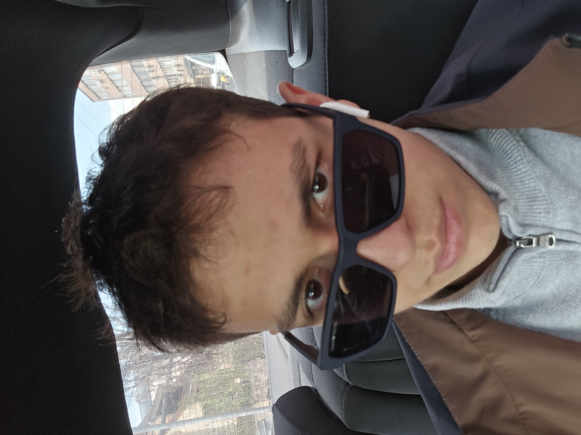 A person wearing sunglasses and a light-gray zippered jacket is in a car. The background suggests the image was taken in Yerevan, Armenia, based on the architectural style visible through the car windows, including a brick building with a distinctive rectangular pattern on its side.
