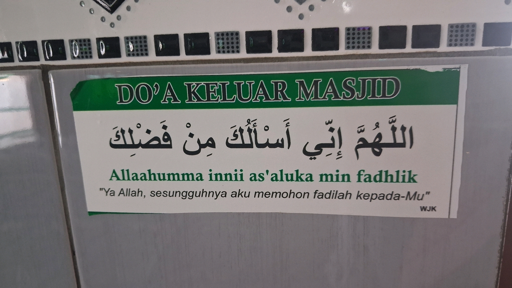 The image displays a sign with religious text in Arabic and Indonesian, likely a prayer for exiting a mosque. The sign is green and white and features the title "DO'A KELUAR MASJID," which translates to "Prayer for Leaving the Mosque." Below the title, in large Arabic script, is the prayer: "اللَّهُمَّ إِنِّي أَسْأَلُكَ مِنْ فَضْلِكَ" (Allahumma innii as'aluka min fadhlika). Underneath the Arabic text, the Indonesian translation is provided: "Allaahumma innii as'aluka min fadhlik" followed by a more colloquial translation in quotation marks: "Ya Allah, sesungguhnya aku memohon fadilah kepada-Mu". A small abbreviation "WJK" is visible in the bottom right corner of the sign. The sign is affixed to a tiled wall, with the tiles appearing to be a light grey or white color. The overall environment suggests a public space, possibly an entrance or exit area of a mosque or a religious building. The specific location of Giriwarno, Indonesia, is not directly observable in the image itself but would be the context provided for this scene.