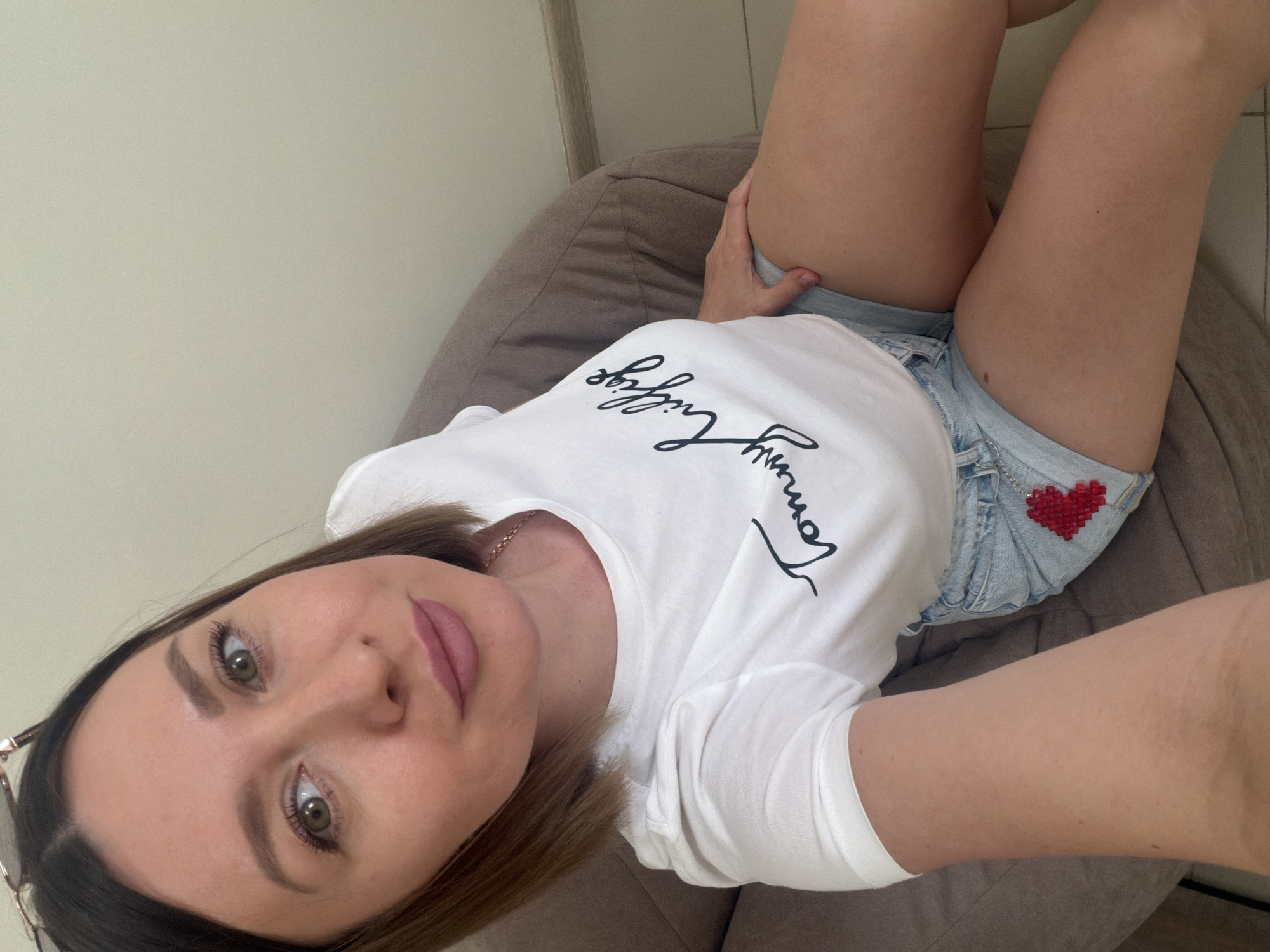 An image taken from an overhead perspective shows a woman reclining on a grey-brown bean bag chair in an indoor setting, likely in Rostov-on-Don, Russia. She has light brown hair, striking green or hazel eyes, and is wearing pink lipstick. Her gaze is directed upwards towards the camera, and her expression is neutral and relaxed.
She is dressed in a white t-shirt with black script text across the chest that clearly reads "Tommy Hilfiger." She also wears light blue denim shorts, to which a small, red beaded heart charm is attached on the right side. A delicate gold chain necklace is visible around her neck, and the frame of a pair of sunglasses can be seen near her left temple. Her right hand is resting on her upper thigh. The background consists of a plain, light-colored wall, indicating an uncluttered environment. The lighting suggests it is daytime.