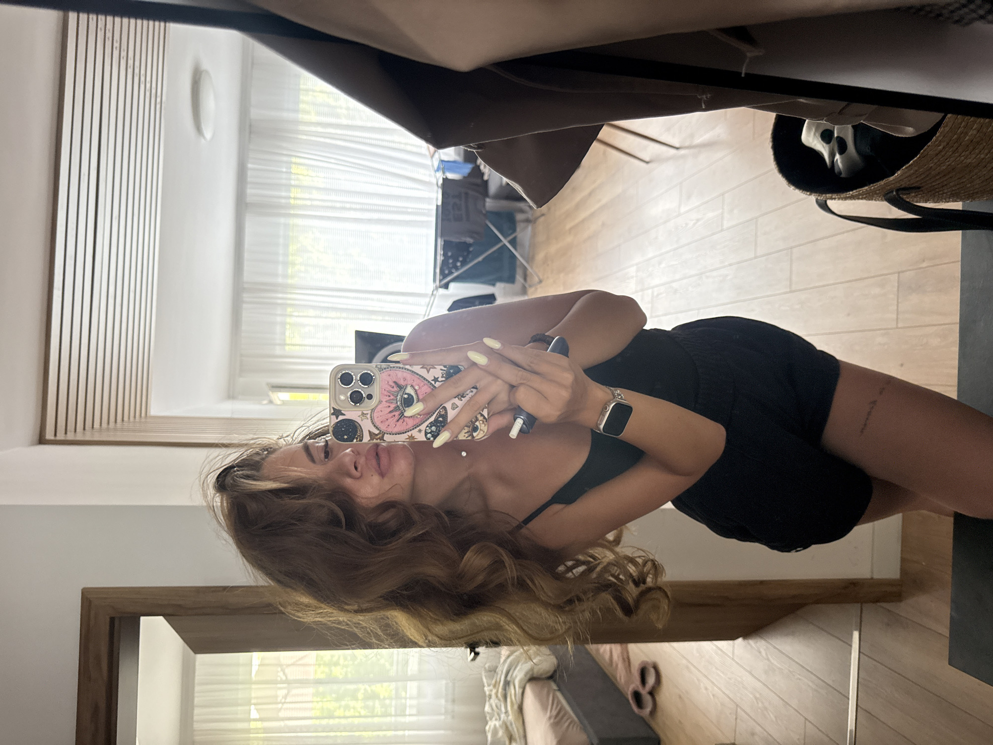 A young woman with long, wavy brown hair is taking a mirror selfie in what appears to be a bedroom or living area. She is wearing a black tank top and black shorts. Her phone, with a pink case featuring an eye design, is held up to take the picture. She is wearing a watch on her left wrist and has light-colored nail polish on her fingernails. In the background, a bed is visible, and through a doorway, a clothes drying rack with laundry can be seen. The flooring is light-colored wood. The overall scene suggests a casual, indoor setting. This image is not geotagged and therefore cannot be definitively placed within the city of Plovdiv, Bulgaria.
