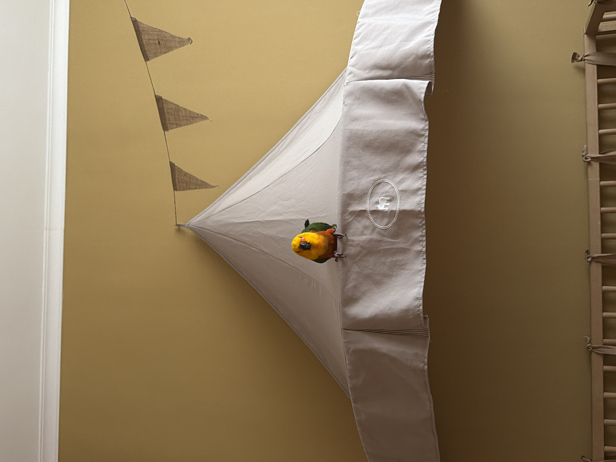 A colorful bird with a yellow head and green and red wings is perched within a triangular off-white fabric structure affixed to a mustard-colored wall. Above the structure, a string of small burlap triangular flags is visible. Adjacent to the fabric structure on the right is a wooden ladder leaning against the wall. To the left of the wall, a white door frame is partially visible. The location information is irrelevant.