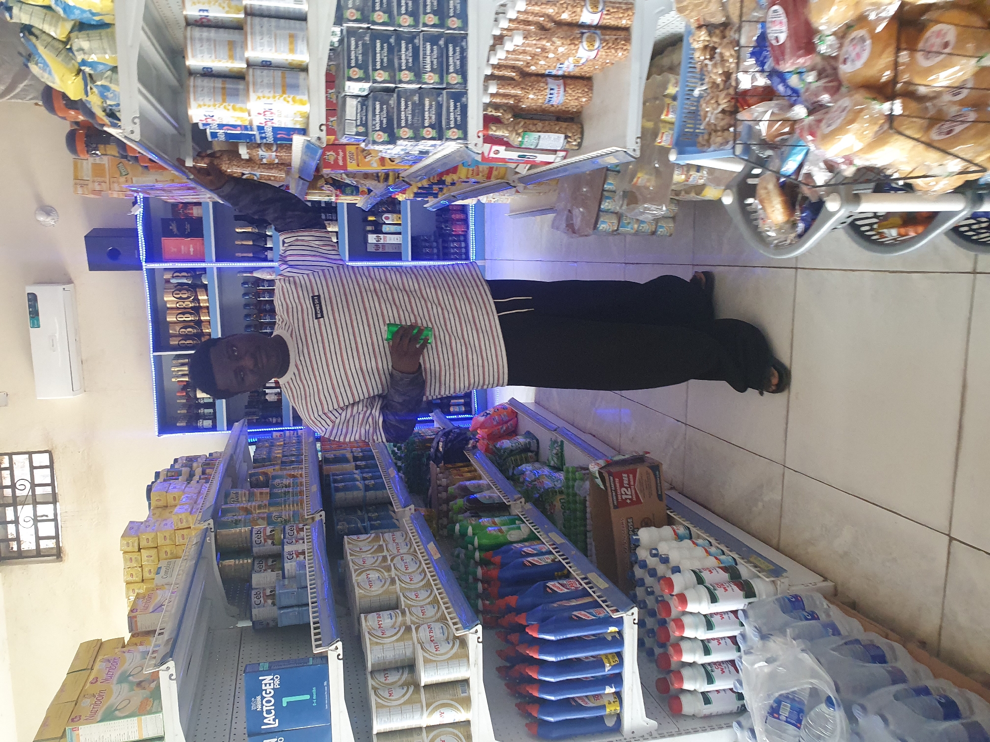 A man stands in an aisle of a store in Anagada, Nigeria. Shelves are stocked with products, including Nutribom, Complan, Lactogen, Ceb, and Golden Penny. Other items visible include bottles of water, cleaning products, and nuts in plastic containers. He is dressed in a striped shirt and dark pants. He is holding a green rectangular object. A shopping cart with bread rolls sits at the end of the aisle. An air conditioner unit is installed on the wall.