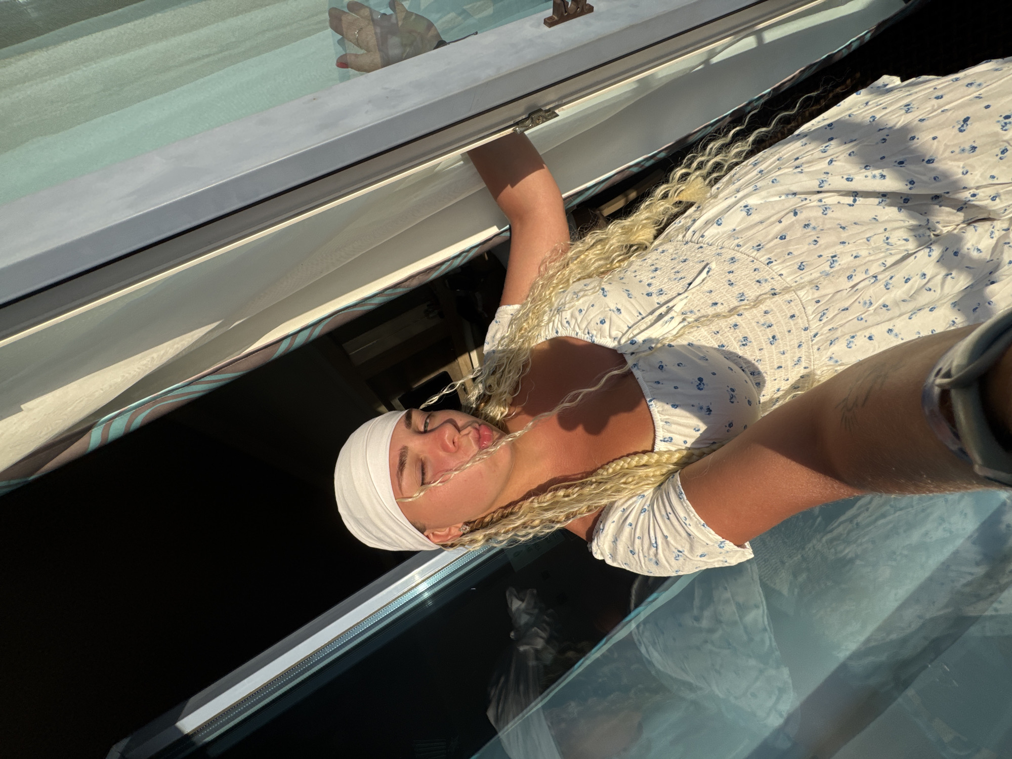A young woman with blonde braided hair and a white headscarf is leaning out of a window, her left arm resting on the window frame. She is wearing a white dress with a blue floral pattern. The sun is casting a warm glow on her face and arm. Her right hand is visible on the glass of the window, appearing to be pushing it open or closed. In the reflection of the window, a person's head and shoulders are visible. The background consists of curtains and a dark interior space. The image appears to be taken from a balcony or terrace overlooking a city, potentially Nesebar, Bulgaria, given the architectural style suggested by the window and railings.