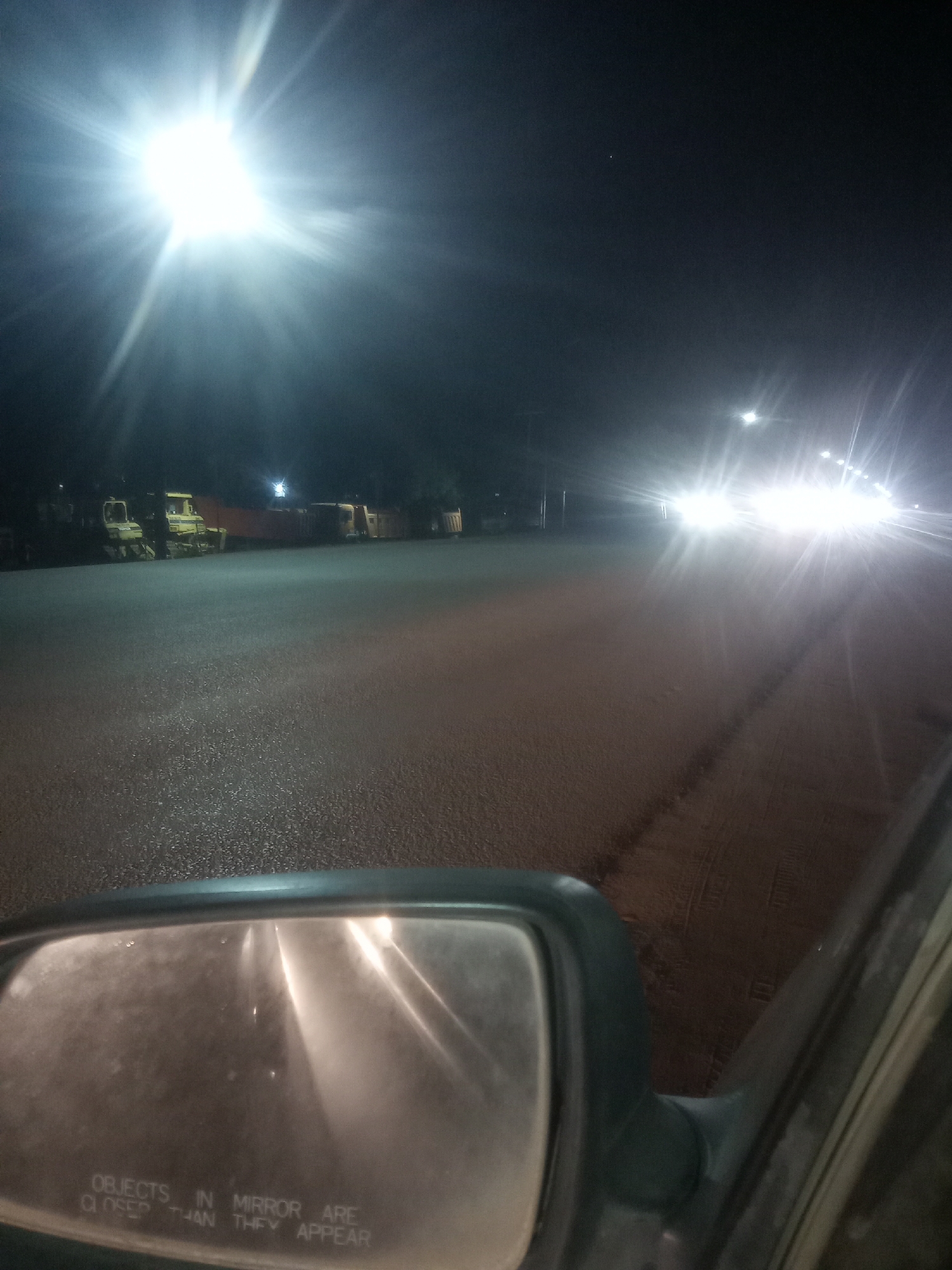 The image depicts a nighttime scene on a road, potentially in Galadimawa, Nigeria. Streetlights illuminate the road surface, which appears wet. Heavy machinery is parked on the side of the road. A side mirror of a vehicle is visible in the foreground, with the text "OBJECTS IN MIRROR ARE CLOSER THAN THEY APPEAR" printed on the mirror's surface. Bright headlights from an approaching vehicle are seen in the distance. The overall environment appears dark and sparsely populated.