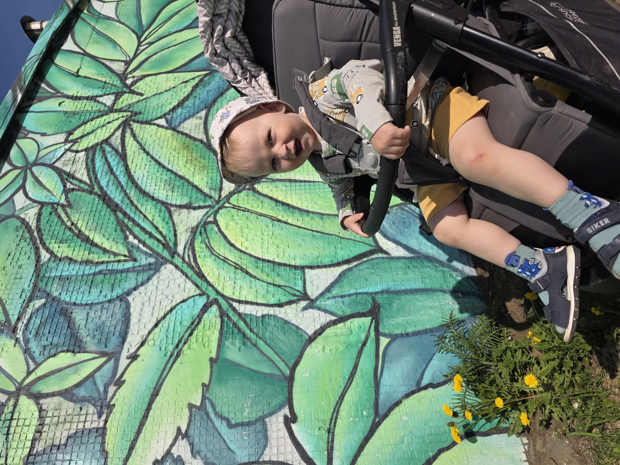 A young child sits in a stroller against a vibrant, green, leafy mural. The child is fair-skinned, wearing a patterned shirt, yellow shorts, a patterned hat, blue socks with vehicle designs, and blue sandals. They are holding onto the stroller's handle and looking at the camera. The stroller is a dark gray color. Yellow dandelions grow near the stroller's wheels. The mural appears on a textured surface and is composed of shades of green. The location is Chaikovsky, Russia.