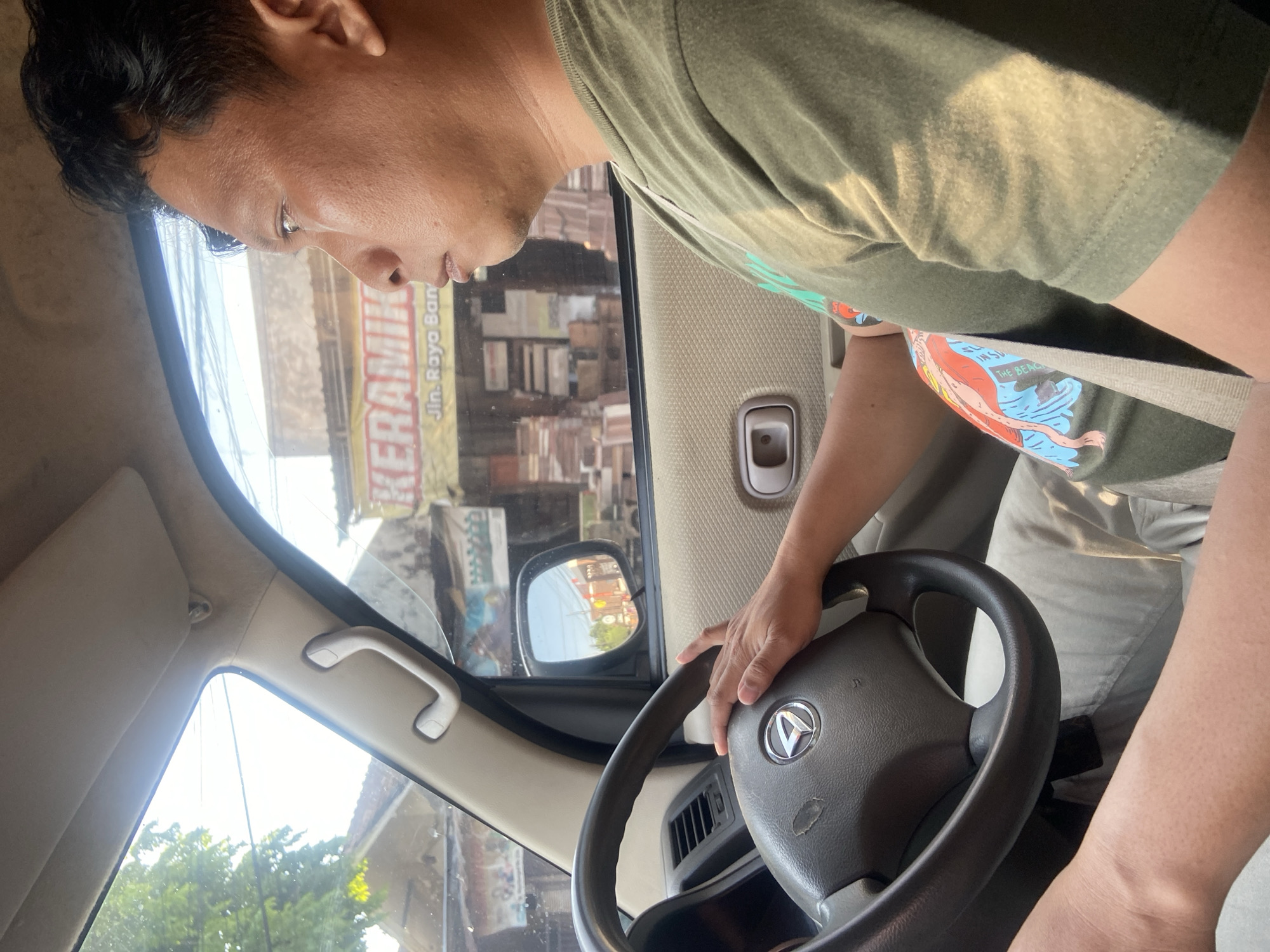A man with dark hair is driving a car on a sunny day. He is wearing a green t-shirt and khaki pants, and a seatbelt is visible. The car's interior is visible, including the steering wheel, dashboard, and a side mirror. Through the side window, a street scene in Bangsal, Indonesia, can be seen, featuring buildings, trees, and signage that reads "KERAMIK" and "Jln. Raya Ban."