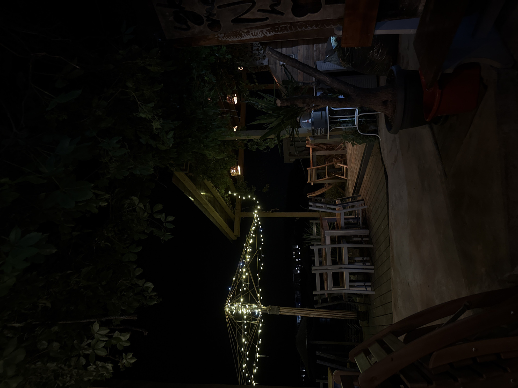 An outdoor seating area of a restaurant or bar is depicted at night. The scene is lit by strings of small, warm-toned fairy lights draped over a large, bamboo-framed umbrella and from an overhead structure. In the foreground, several wooden chairs are stacked or leaning against each other. Behind them, a wooden deck is visible, furnished with tables and chairs, suggesting a dining or seating arrangement.
Further back, a plant in a large pot stands near some wooden structures and what appears to be a large trash bag. A sign with dark lettering is visible on the right side of the image. The background is dark, with a few distant lights suggesting a town or settlement along a waterfront. The overall atmosphere is relaxed and atmospheric, characteristic of a seaside establishment in the evening. The location is identified as Tsarevo, Bulgaria.