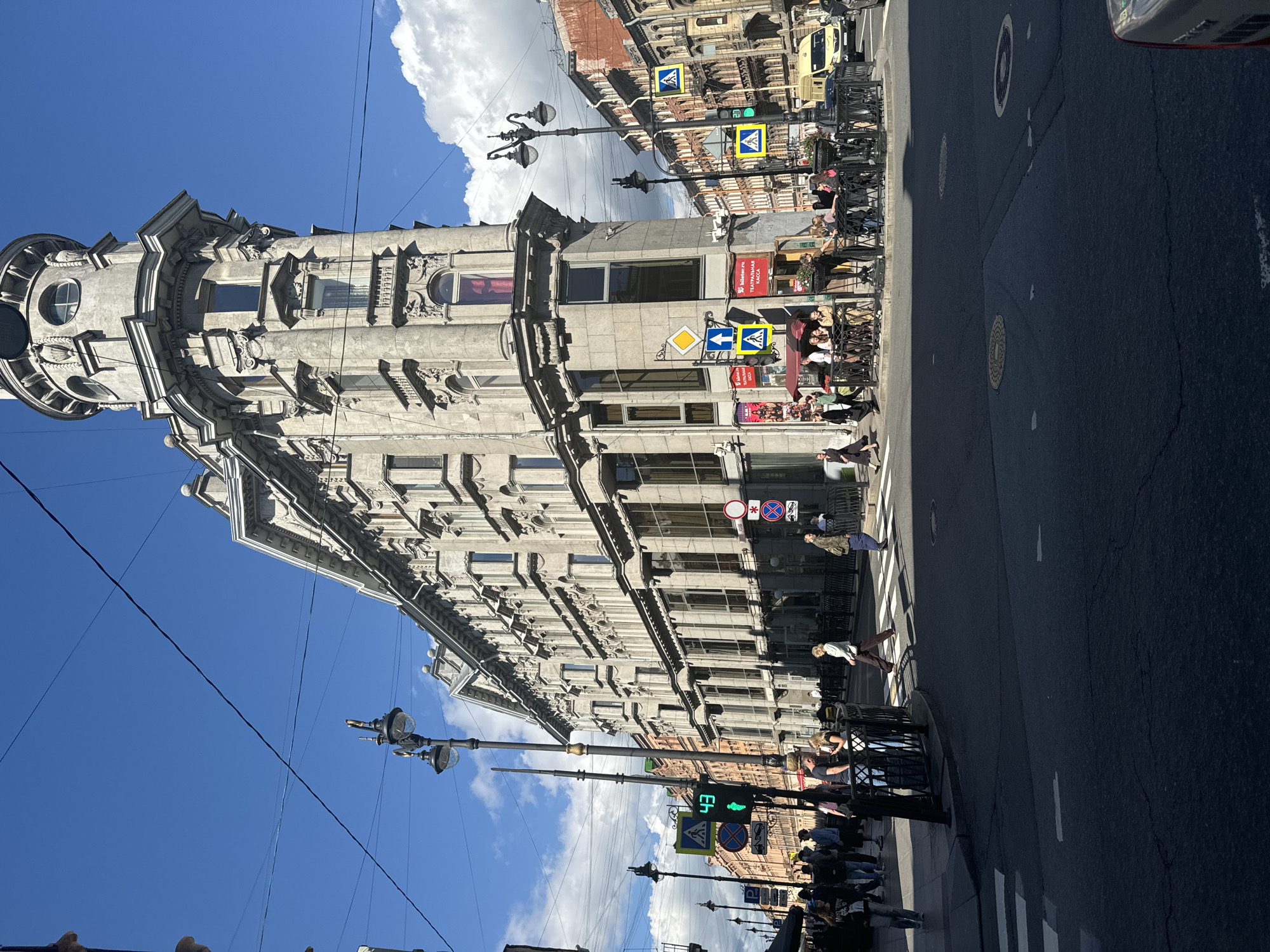 The image depicts a street intersection in Saint Petersburg, Russia, dominated by a distinctive triangular building with intricate architectural details. The building, a pale gray stone structure, features multiple stories, ornate balconies, and a rounded corner tower with circular windows and decorative elements. It is situated on a sunny day with a bright blue sky and scattered white clouds.
In the foreground, a crosswalk is visible, with several pedestrians in the process of crossing or waiting to cross. The people are dressed in casual summer attire. A traffic light with a green signal for pedestrians is prominent on the left side of the intersection. Other street furniture includes lampposts with multiple lights and various street signs, including pedestrian crossing signs and traffic regulations.
To the right of the triangular building, other buildings with orange and gray facades line the street. A yellow van is parked near the intersection, and streetlights are visible throughout the scene. The street is paved with asphalt, and manhole covers are present on the road. The overall impression is of a bustling urban environment on a pleasant day, capturing a slice of daily life in Saint Petersburg.