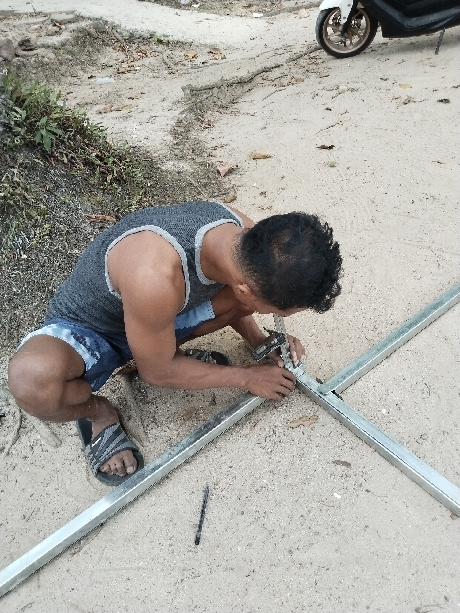 In Prigi, Indonesia, a man wearing a grey tank top and blue and white patterned shorts kneels on sandy ground, working on a metallic frame. He is holding a caliper to measure a joint in the metal structure. A black pen lies on the ground nearby. In the background, a motorcycle is partially visible, and the ground is covered in sand, dry leaves, and some sparse vegetation.