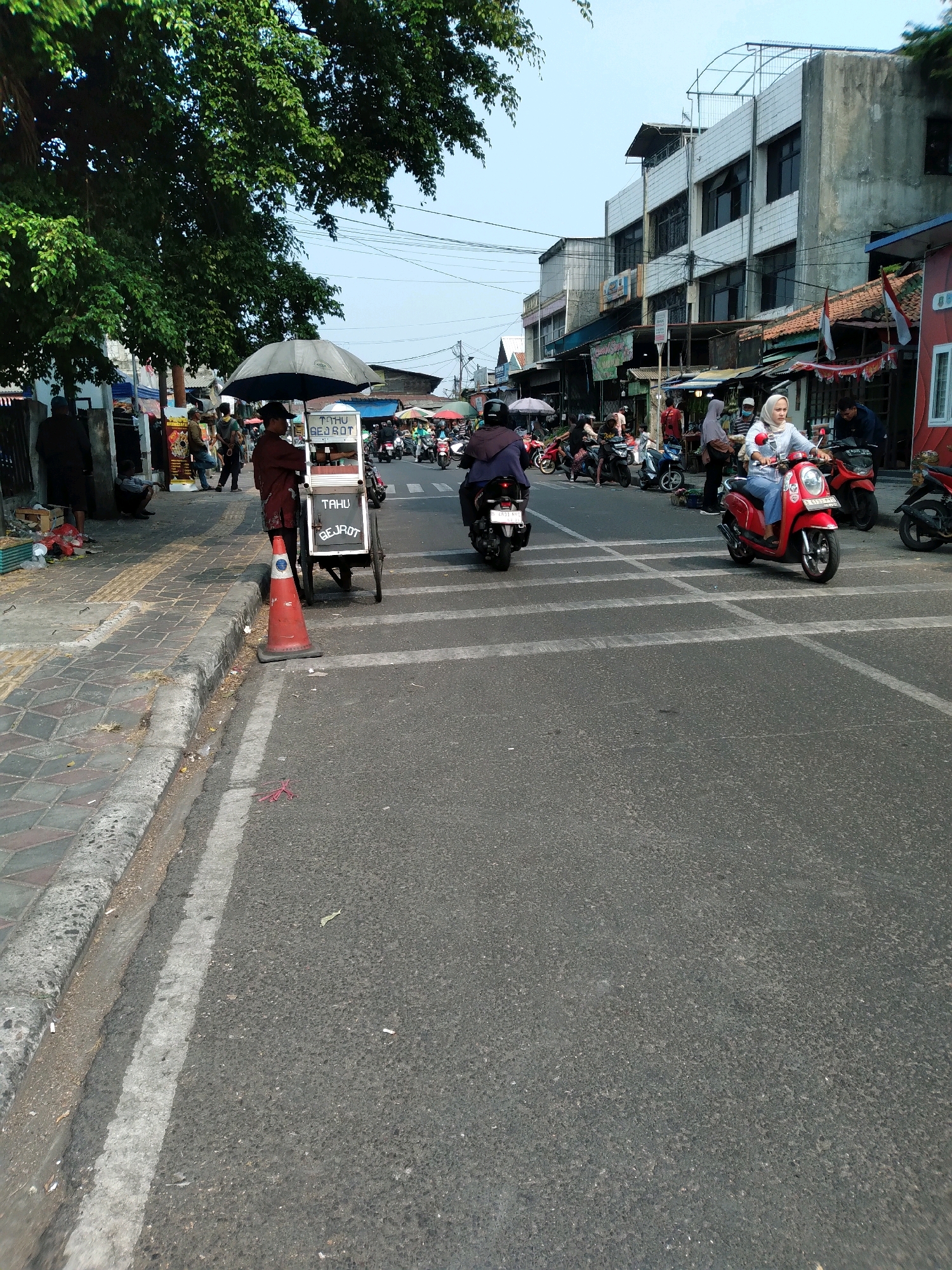 The image depicts a street scene in South Tangerang, Indonesia, likely during daylight hours given the bright illumination and clear sky. The foreground shows a paved road with white lane markings. On the left side of the road, a pedestrian walkway with patterned tiles is visible, separated from the road by a curb. A street vendor's cart, topped with an umbrella and displaying the text "TAHU BEJROT", is parked on the walkway. An orange traffic cone is placed near the cart.
Several people are present in the scene. A vendor stands behind the cart. Further down the walkway, other individuals are seen near market stalls or shops. On the road, a woman wearing a hijab and blue jeans rides a red scooter, while another person on a motorcycle is visible further back, as is a third person on a scooter. In the background, buildings line the street, some appearing to be multi-story commercial or residential structures. The overall atmosphere suggests a busy urban environment with ongoing commercial activity.
