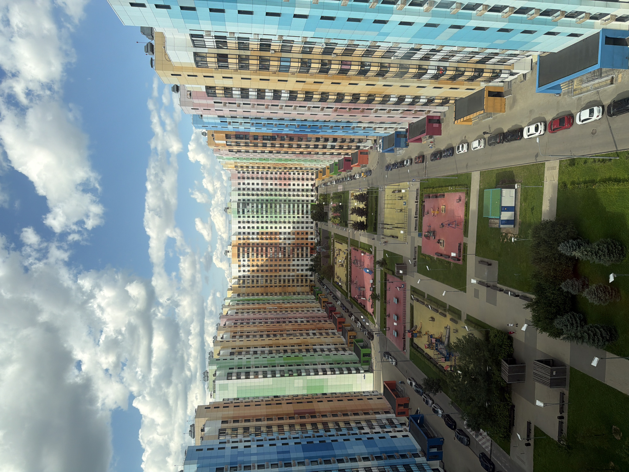 This is an aerial, eye-level, full shot of a modern, colorful residential complex with multiple tall buildings of varying bright hues under a partially cloudy sky. The buildings are arranged in rows, creating a long, colorful corridor. In the foreground, a paved walkway is bordered by manicured green lawns and trees. Several children's playgrounds with red and yellow surfaces are visible, equipped with swings, slides, and climbing structures. A road runs alongside the buildings, with a line of parked cars, mostly dark-colored sedans, and a few SUVs. Some of these cars are parked on the street, while others are in designated parking areas. There are also a few people walking on the sidewalks and in the courtyards. Small, brightly colored structures are situated at the base of some buildings, possibly entrances or utility rooms. The buildings themselves feature numerous windows and balconies, indicating residential units. The overall impression is one of a well-maintained, contemporary urban living space.