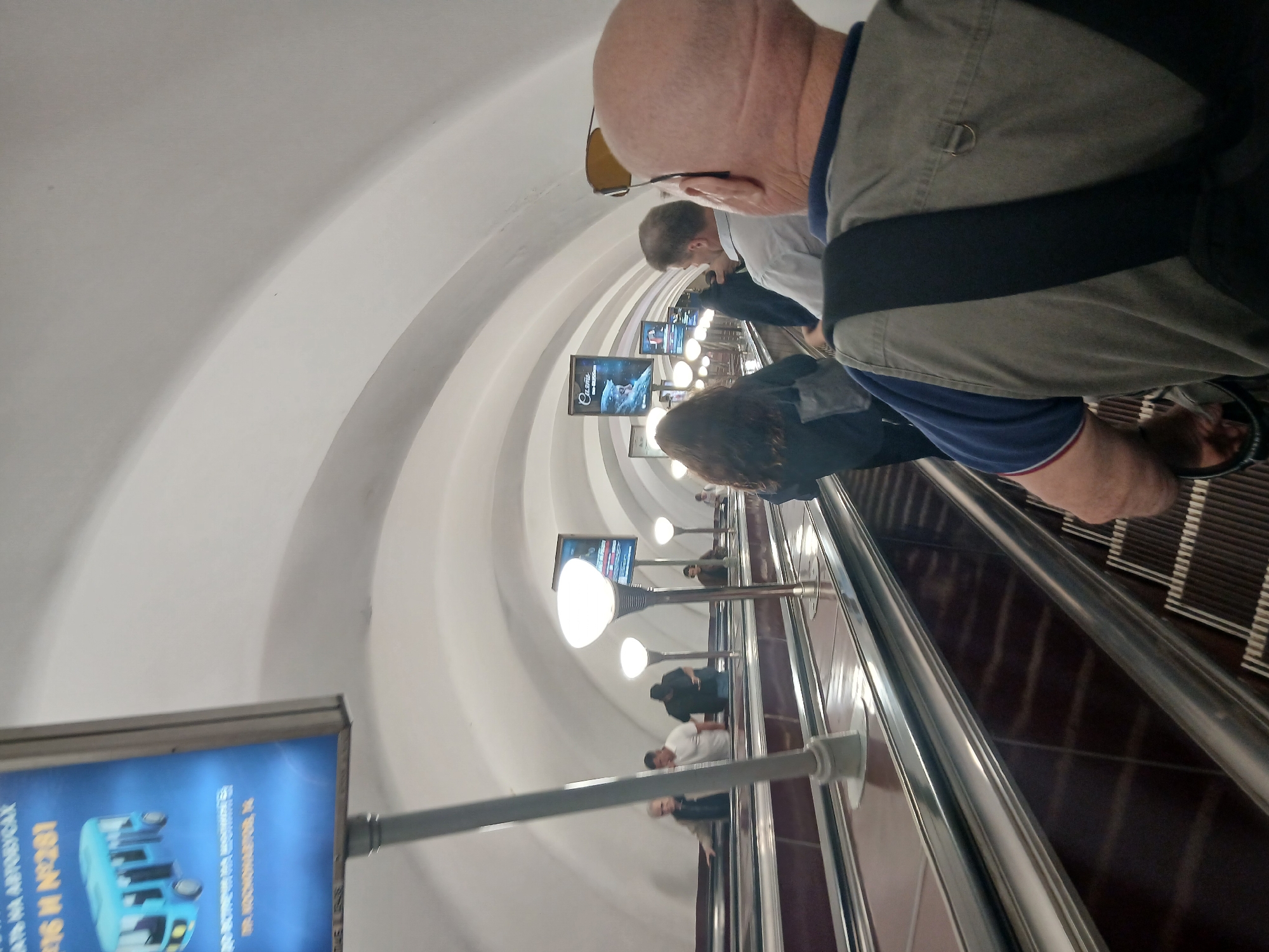 An outdoor shot captures a group of people on an escalator, ascending into a white, arched ceiling, lit by intermittent lamps. In the foreground, a bald man with sunglasses on his head wears a blue collared shirt with a khaki vest over it. To his left, a woman with long, dark hair is also ascending. Further up the escalator, several other individuals are visible, some walking and others standing. The escalator itself is lined with metal and dark-colored material. A blue sign with a drawing of a bus and text in a foreign language is visible on the left side of the image. The scene appears to be in a public transit area.