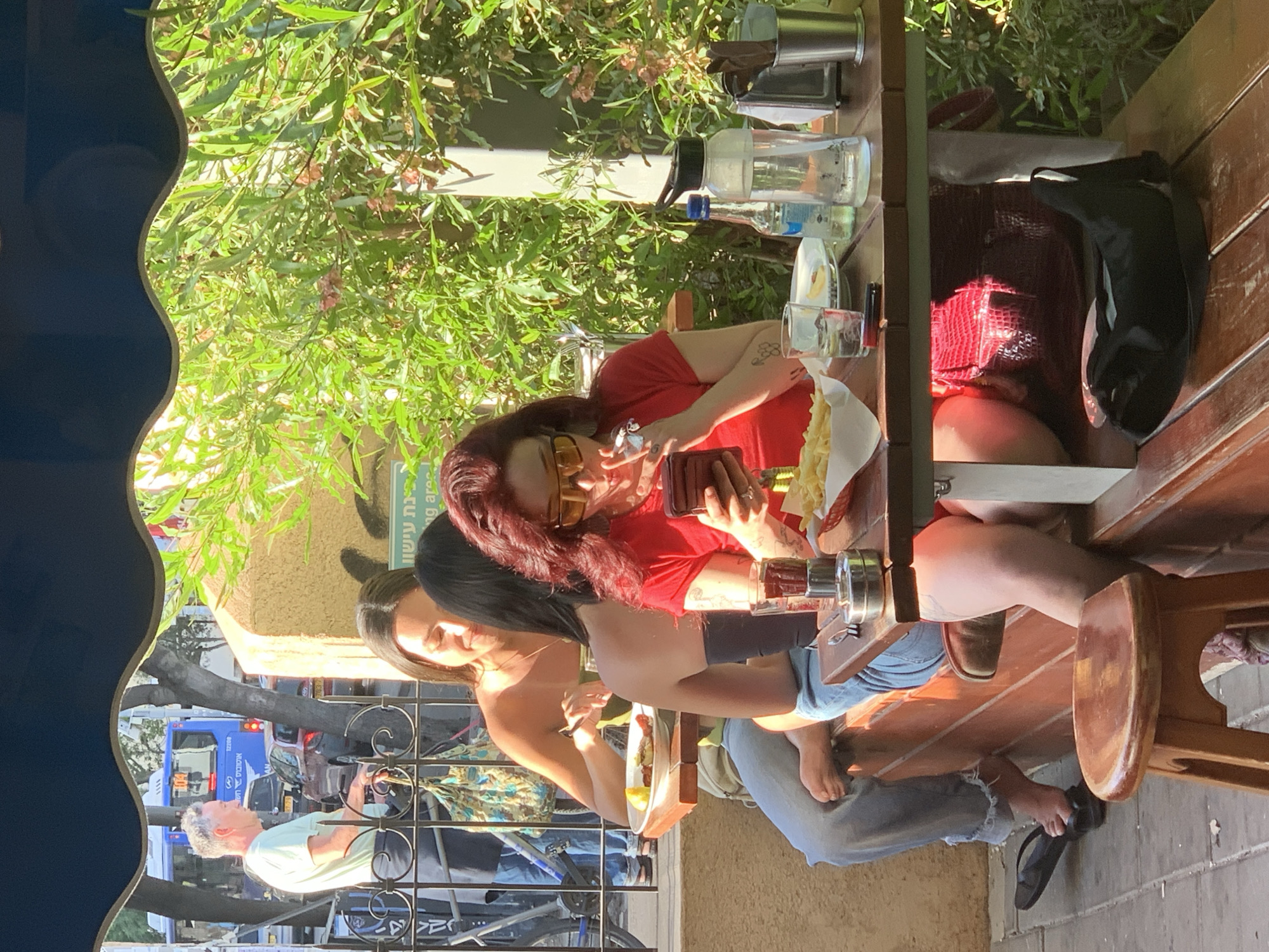 The image captures a vibrant daytime scene at an outdoor cafe or restaurant in Tel Aviv. Three women are seated at a wooden table, enjoying food and drinks in a relaxed setting. The woman closest to the foreground has auburn hair, wears a red top and yellow sunglasses, and is looking at her phone. Across from her, another woman with dark hair, wearing a black top, is also seen interacting with a phone and eating. A third woman, partially visible to the left, wears a light-colored top.
On the table, various items are present, including multiple glasses of drinks, water bottles, and a large bowl of what appears to be fries or pasta. One woman has a plate of food, possibly grilled items, in front of her. Green foliage and trees provide a natural, shaded canopy over the dining area.
Beyond the immediate cafe setting, an urban street is visible in the background, bustling with traffic. A blue Metropoline public bus, bearing the Hebrew text "מטרופולין" (Metropoline) and "קו 82239" (Line 82239), is prominent. Other cars and a man in a light green polo shirt can also be seen on the street. A black metal fence separates the cafe from the sidewalk and street.
The scene suggests a sunny day, with bright light filtering through the trees. The overall mood appears casual and social. Visible Hebrew text also includes "בתיאבון" (Bete'avon, meaning Bon appétit or Enjoy your meal) on a green sign in the background. One of the women at the table is barefoot, with her feet resting on a lower wooden beam of the table.