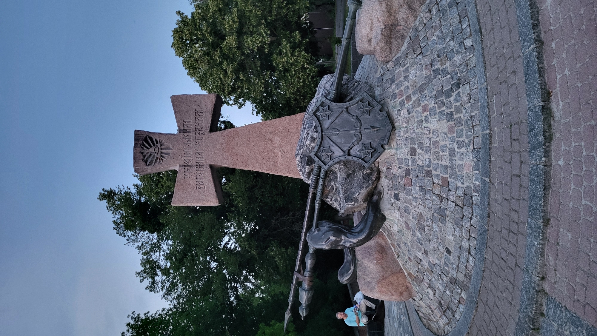 The image depicts a monument with a large, reddish-brown cross as its central feature. The cross is adorned with carved inscriptions and a sun-like symbol. To the right of the cross, a metallic shield with a star pattern is mounted on a large stone. Below the shield, a spear and a sword are part of a sculpture, possibly representing a warrior or a historical figure. The monument is situated on a cobblestone pavement, with some large boulders incorporated into its base. In the background, lush green trees are visible against a twilight sky. Two people are seated on a low wall or bench in the lower right of the frame, seemingly resting or observing the monument. The overall scene suggests a public space, possibly a memorial or a historical site. The lighting indicates it is late evening or early morning. The inscription on the cross is in Cyrillic script and reads "ВЕЛИКОМУЧЕНИКЪ ЗАСТУПНИКЪ РУСИ" which translates to "Great Martyr, Protector of Rus'".