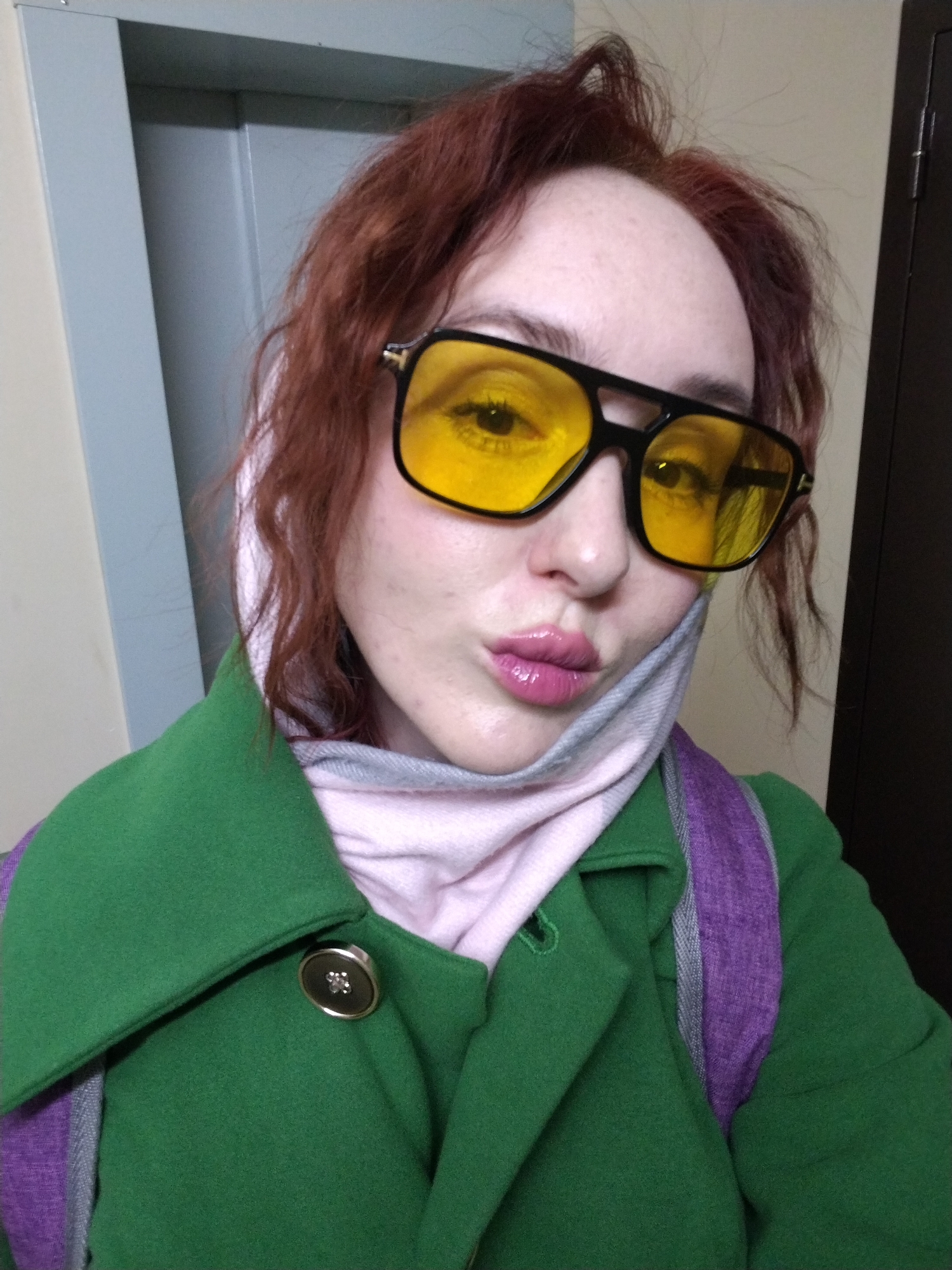 A person, likely female, is captured in a close-up selfie from the chest up. They have reddish-brown, slightly disheveled hair and wear large, black-framed sunglasses with distinct bright yellow lenses, obscuring their eyes. Their lips are pursed in a "duck face" pose and are covered in glossy pink lipstick.
The subject is wearing a vibrant green coat or jacket. A decorative silver-colored button with a subtle patterned design (possibly a starburst or flower) is visible on the coat's lapel on their left side. Around their neck, a light pink and light grey striped scarf is wrapped, providing a soft contrast to the green coat. A purple strap, consistent with a backpack, is visible over their right shoulder.
The background is a simple indoor setting. To the left, a light beige wall meets a grey vertical panel, which could be part of a doorframe or wall trim. To the right, a dark brown door or doorframe is visible. The lighting appears to be artificial, suggesting an indoor environment, with no discernible clues about the time of day or external weather conditions. The overall expression, characterized by the pursed lips and bold glasses, conveys a playful or confident mood. No text is visible in the image.