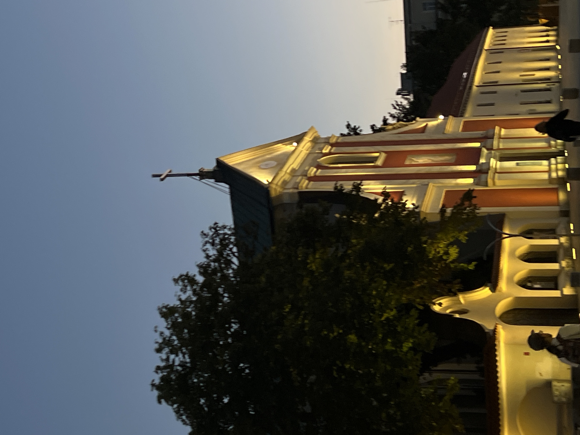 The image depicts the Saint Bernardine Church and Monastery in Lublin, Poland, during twilight. The church's facade, painted red and white with yellow illumination, is prominently featured. A cross stands atop the church's roof. To the left, a large, dark tree partially obscures the view of the church. In the foreground, two individuals are visible. One, on the left, is wearing a cap and backpack, facing away from the camera. Another person, on the right, is walking away from the camera. The sky above is a clear, fading blue. Yellow lights illuminate the building's architectural details and a section of a wall with arched openings.