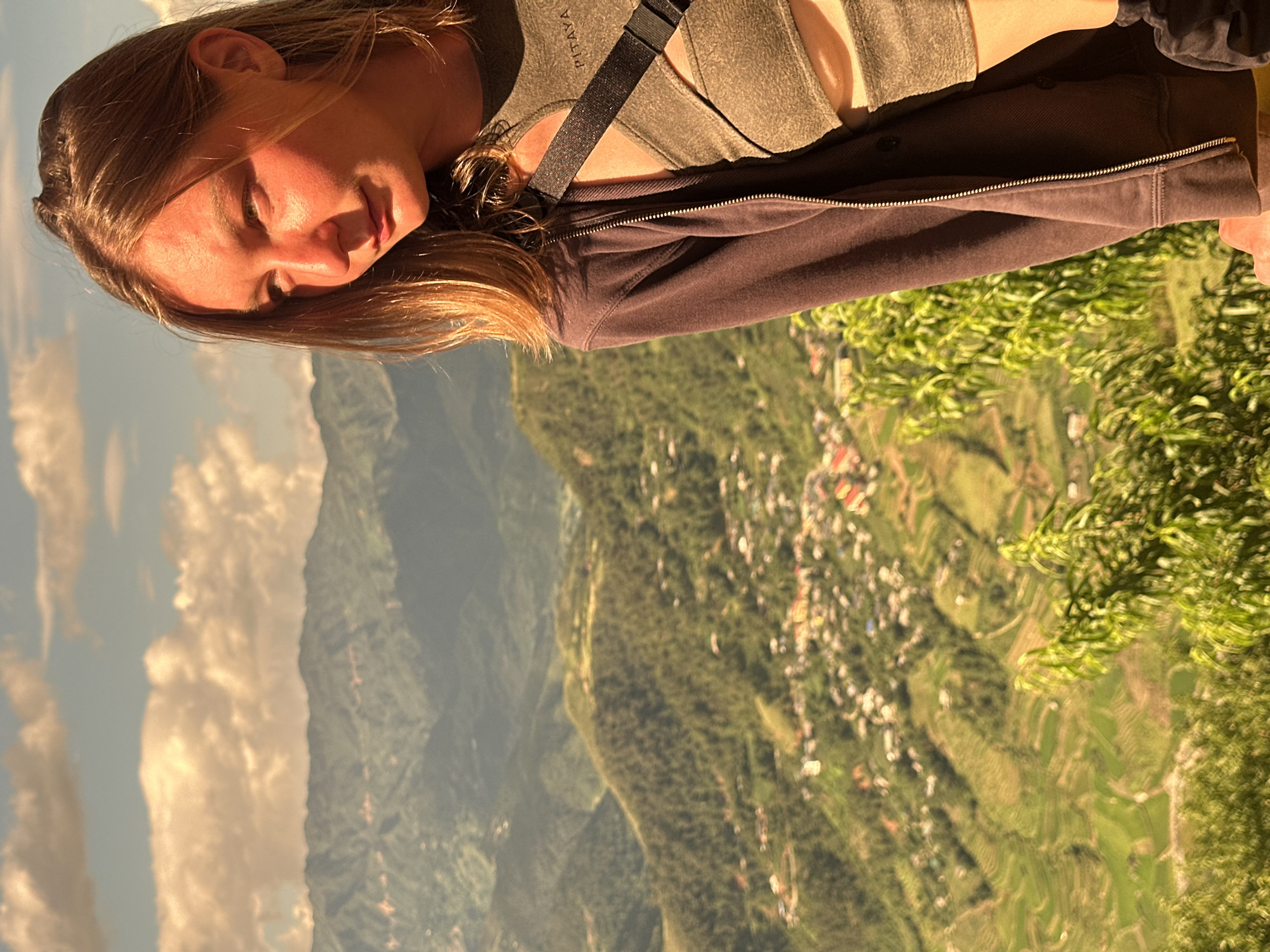 An outdoor, eye-level shot captures a young adult female positioned in the right foreground, looking downwards and to her left, with her profile partially illuminated by strong golden light. She has light brown hair and fair skin, with a warm glow on her right cheek and chin. She is wearing an olive-green textured crop top with cut-outs and a criss-cross design, featuring a faint "PITAYA" logo on the upper chest. A dark, wide strap crosses her chest, and she wears a dark brown/charcoal zippered hooded jacket.
The background reveals a vast mountainous landscape, characteristic of Tu Le, Vietnam. Multiple layers of green, forested mountains extend into the distance under a sky with scattered, warm-toned clouds. A village or town, consisting of numerous buildings with light-colored or red roofs, is visible on the lower slopes and in the valley. Distinct green rice terraces are identifiable on the lower mountain slopes, particularly towards the bottom left and center of the frame. Lush green foliage, including leafy branches, partially obscures the immediate foreground on the right. The scene is illuminated by strong, directional sunlight, suggesting late afternoon or early morning.