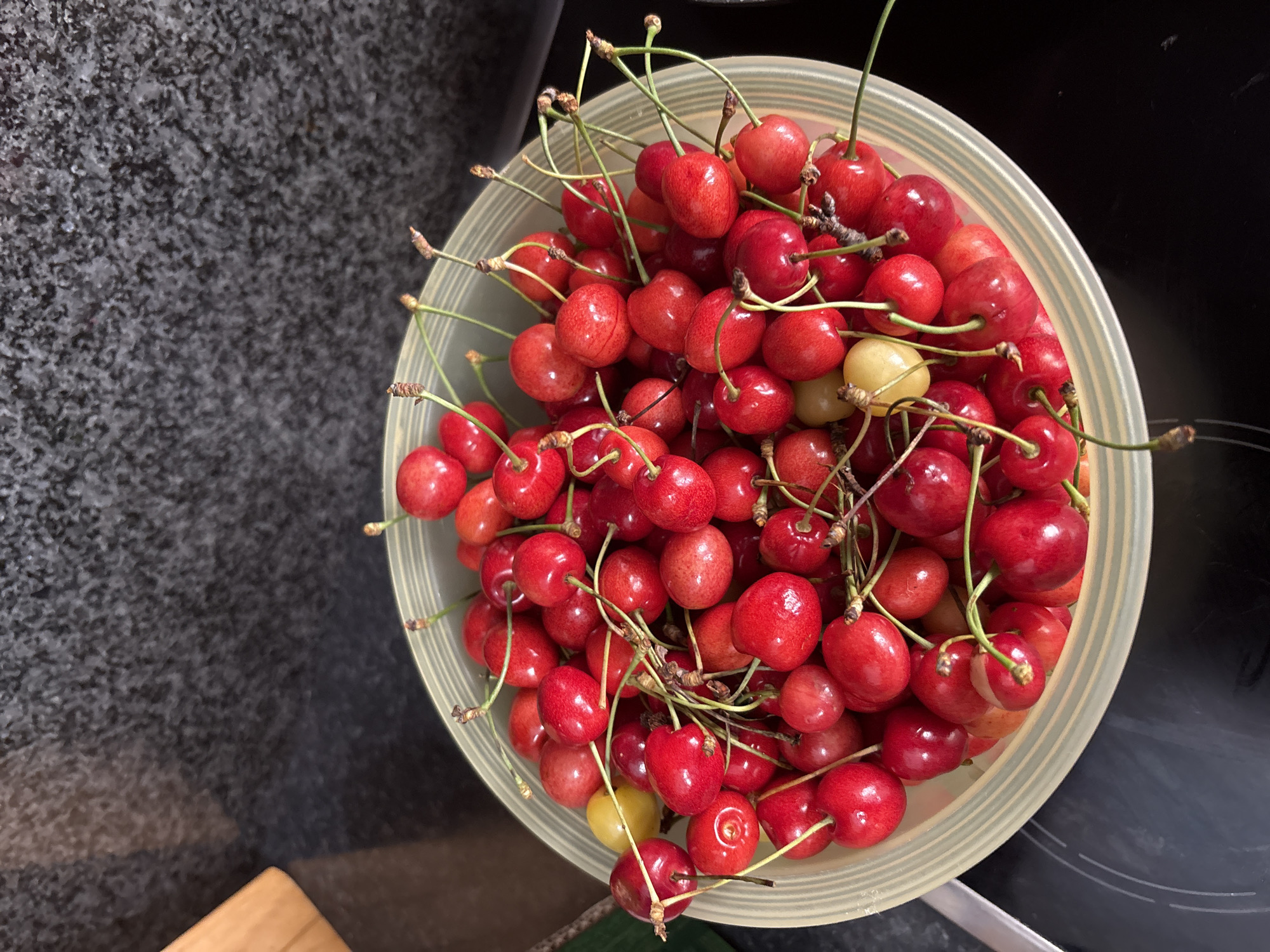 A bowl of fresh cherries sits on a dark, speckled countertop next to a wooden cutting board. The bowl is light green with a ribbed texture and is overflowing with bright red cherries, some with stems still attached. There is also a single yellow cherry in the bowl. The countertop appears to be granite or a similar material. The background is out of focus, showing a dark, textured surface that could be a wall or backsplash. The lighting is bright, casting shadows from the cherries. The image is taken from a high-angle perspective. The scene is located in Ilion, Greece.